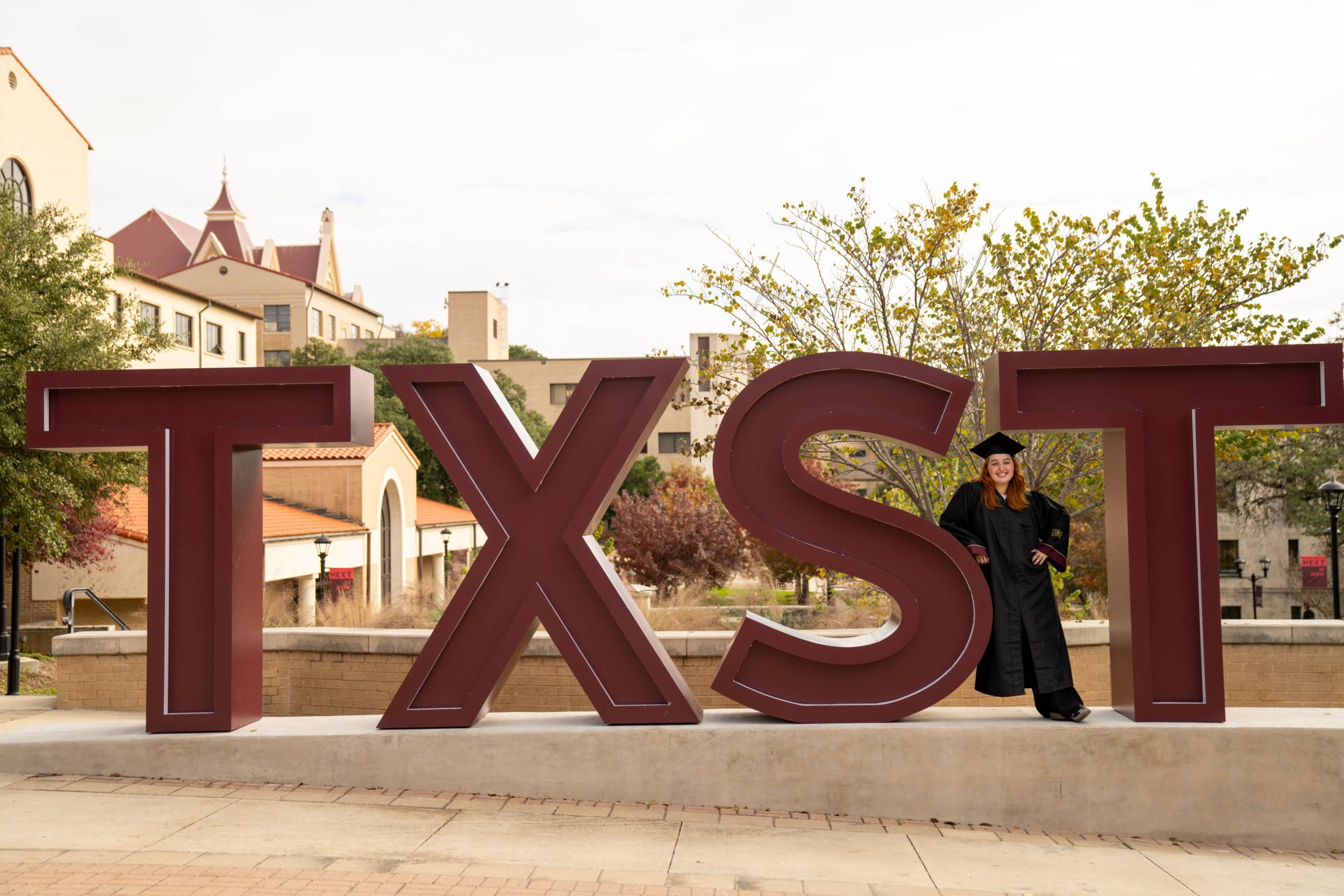 Graduate in cap and gown stands between the large maroon TXST letters on Bobcat Trail, smiling at the camera with campus buildings visible in the background.