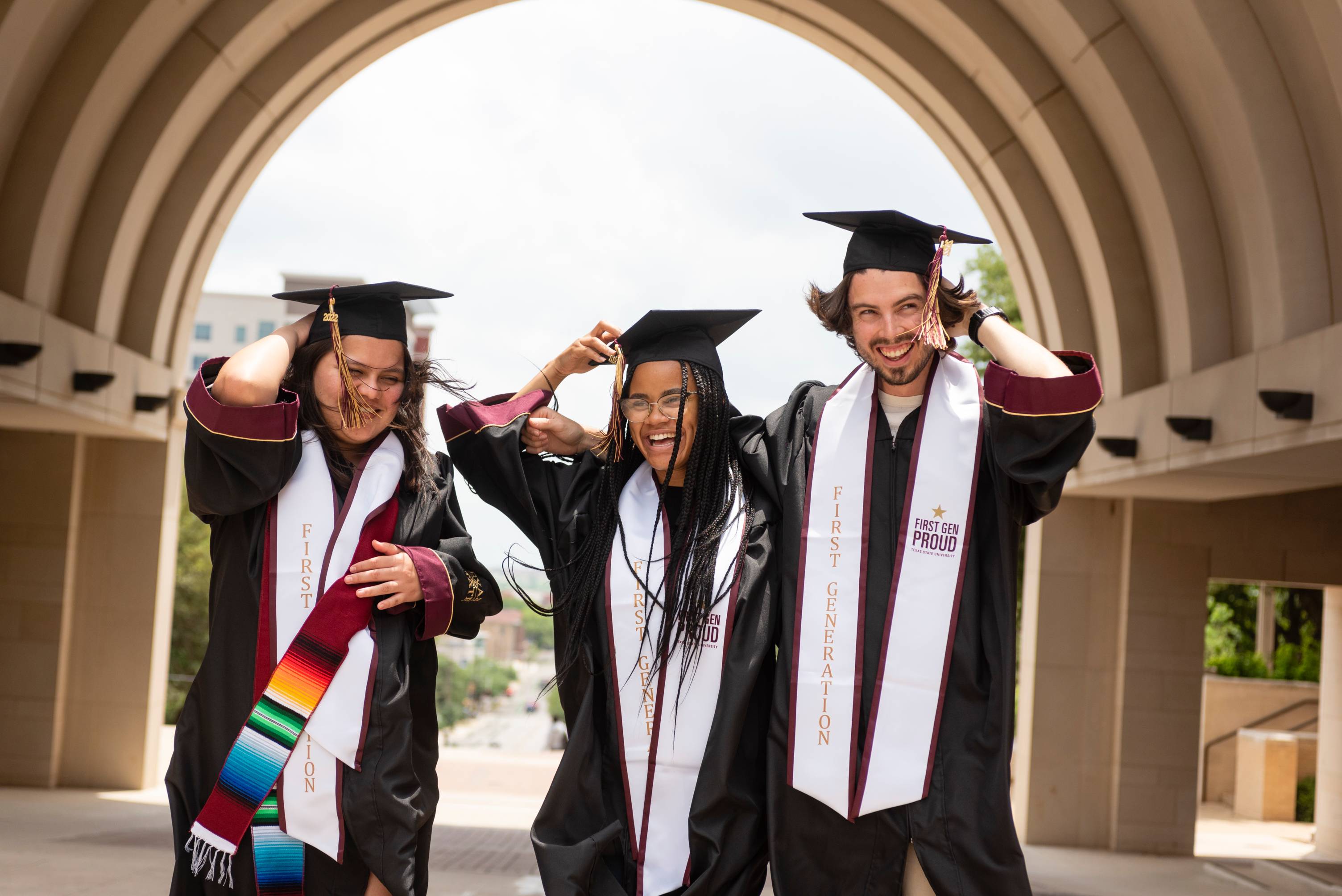 3 graduate in first gen stoles smile and laugh together beneath the UAC Arch.