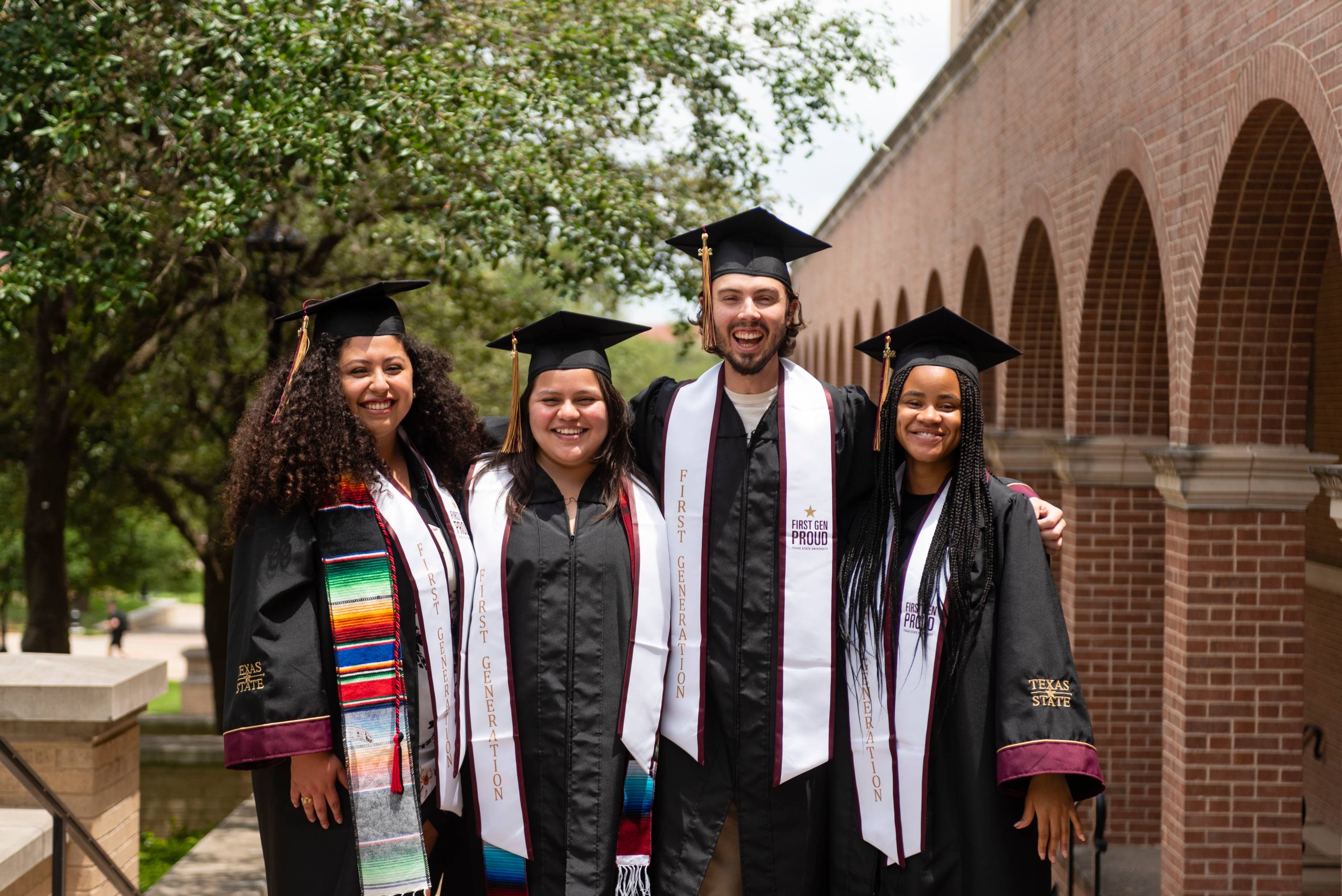 Four graduates in caps and gowns stand together smiling in front of the brick arches at Trauth Huffman, several wearing First Generation stoles.