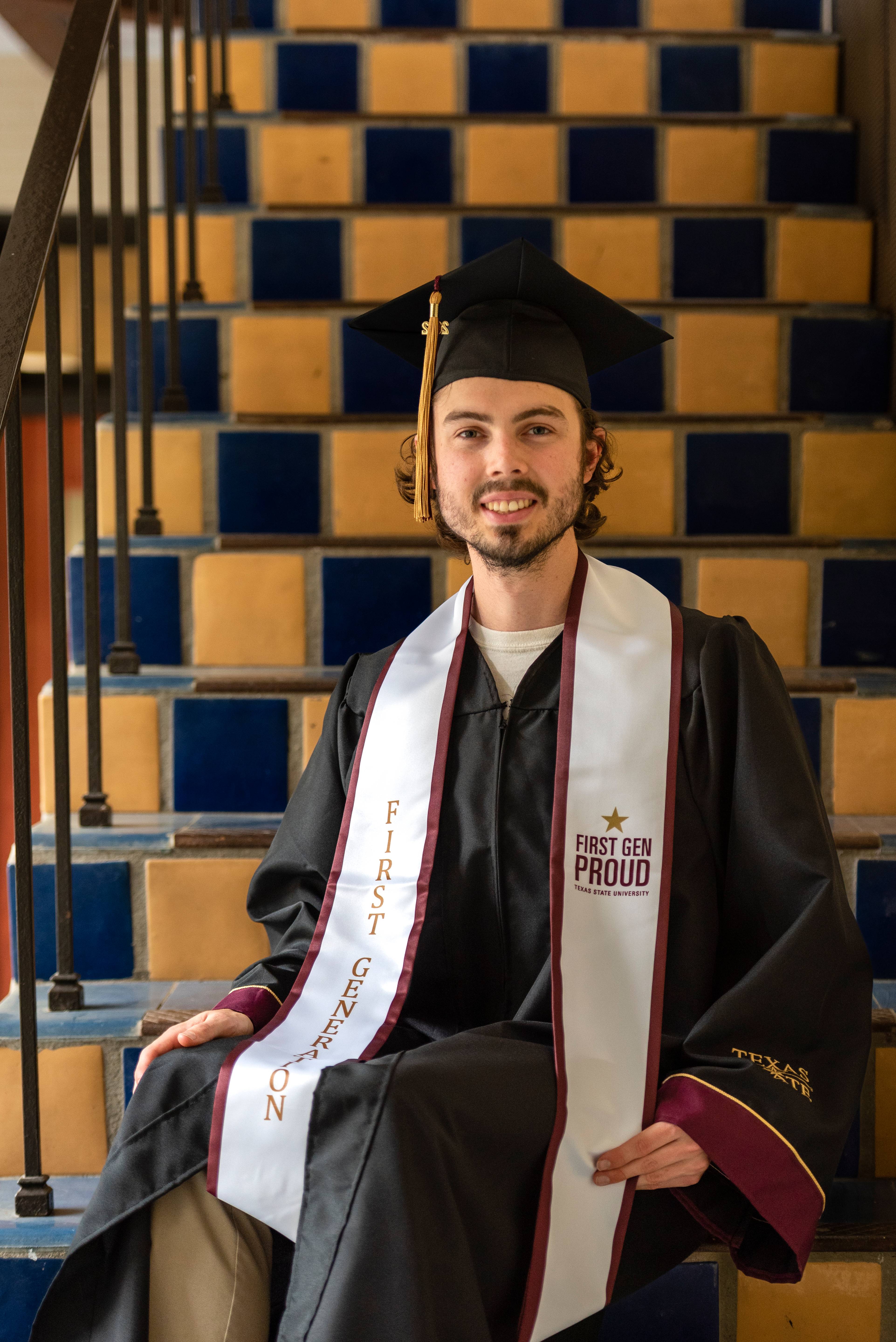 Graduate wearing a First Generation stole smiles at the camera in front of the blue and gold checkered tile wall at Taylor-Murphy Courtyard.