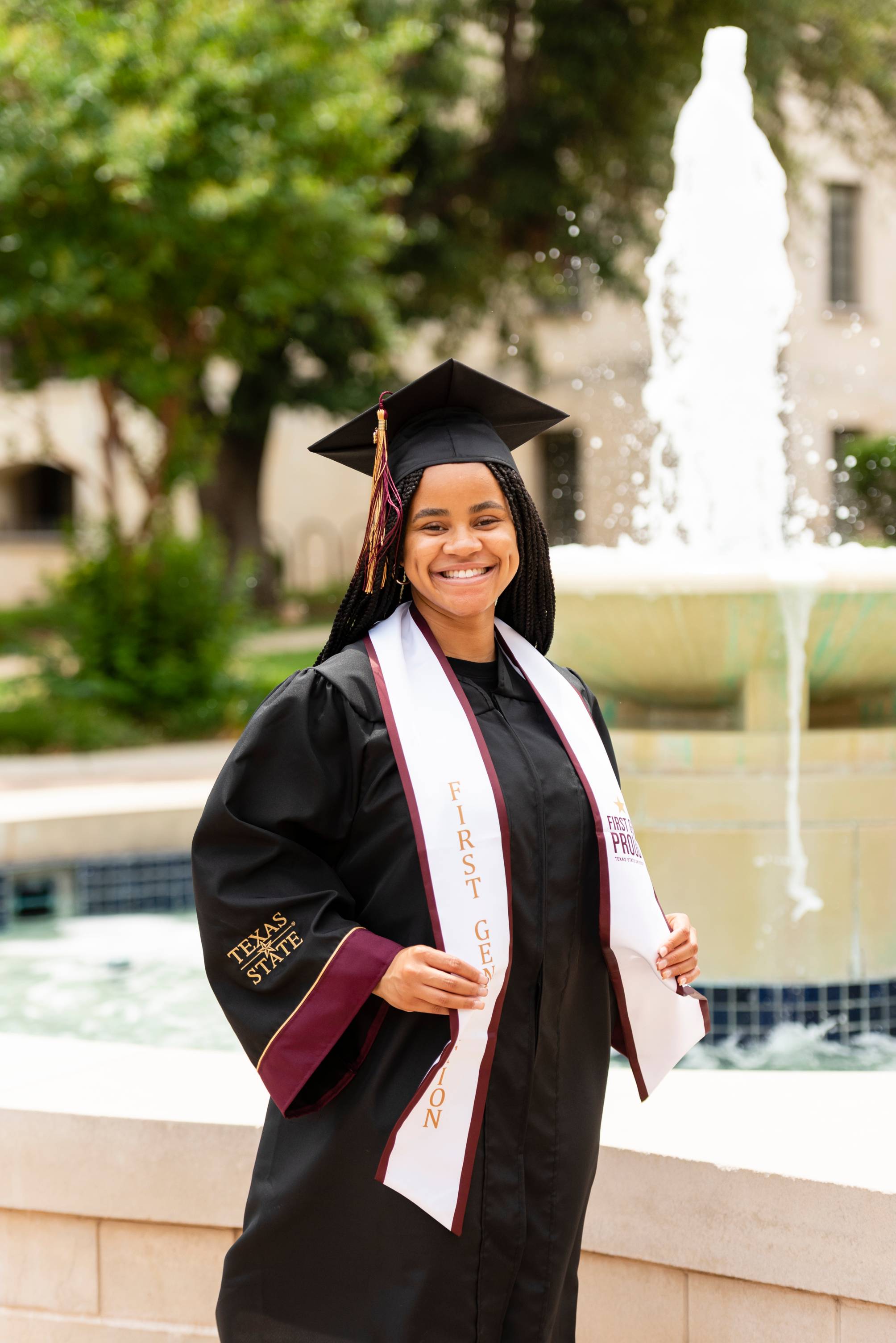 Graduate smiles at the camera in front of the Bobcat Trail fountain.
