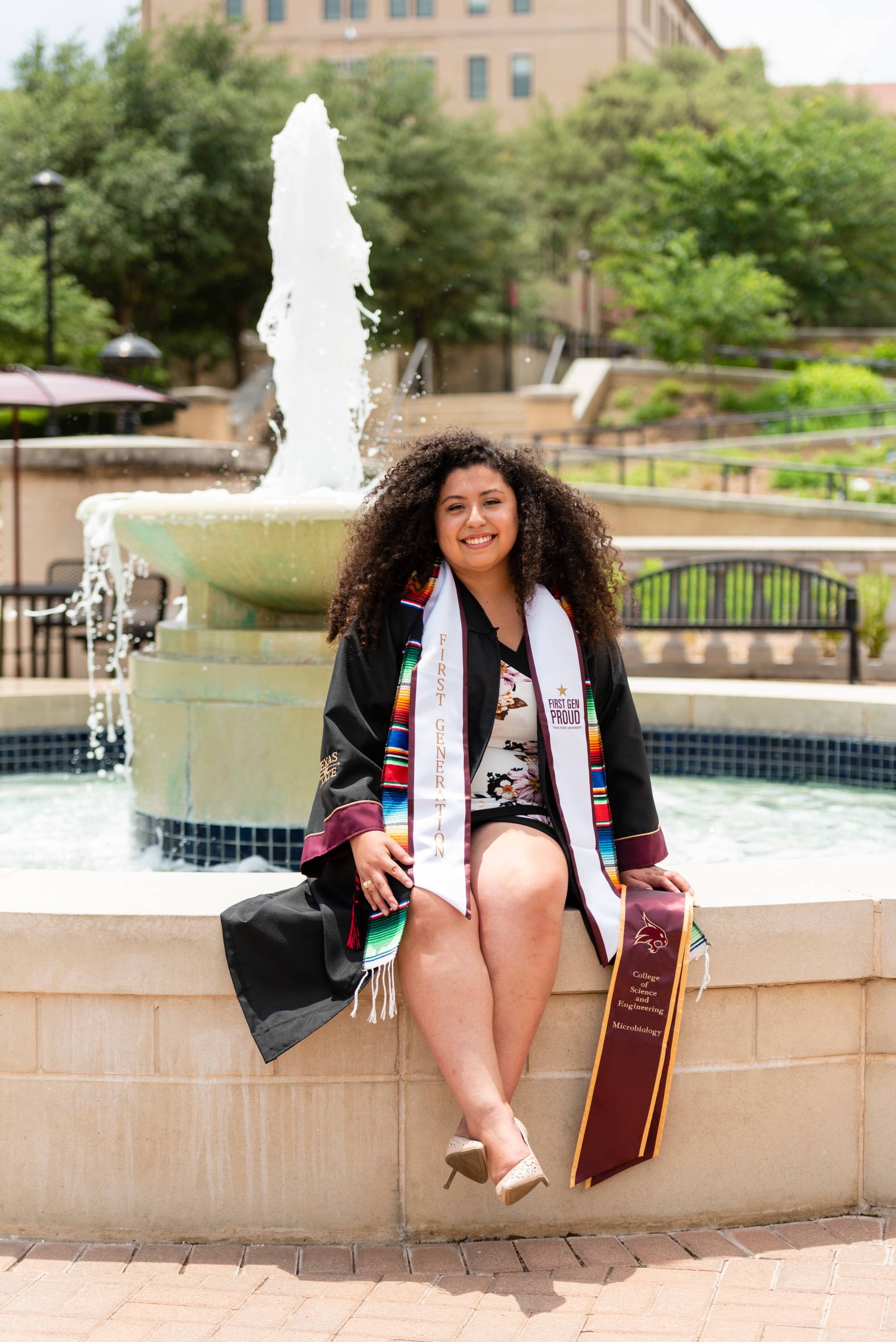 Graduate smiles at the camera sitting on the edge of the Bobcat Trail fountain.
