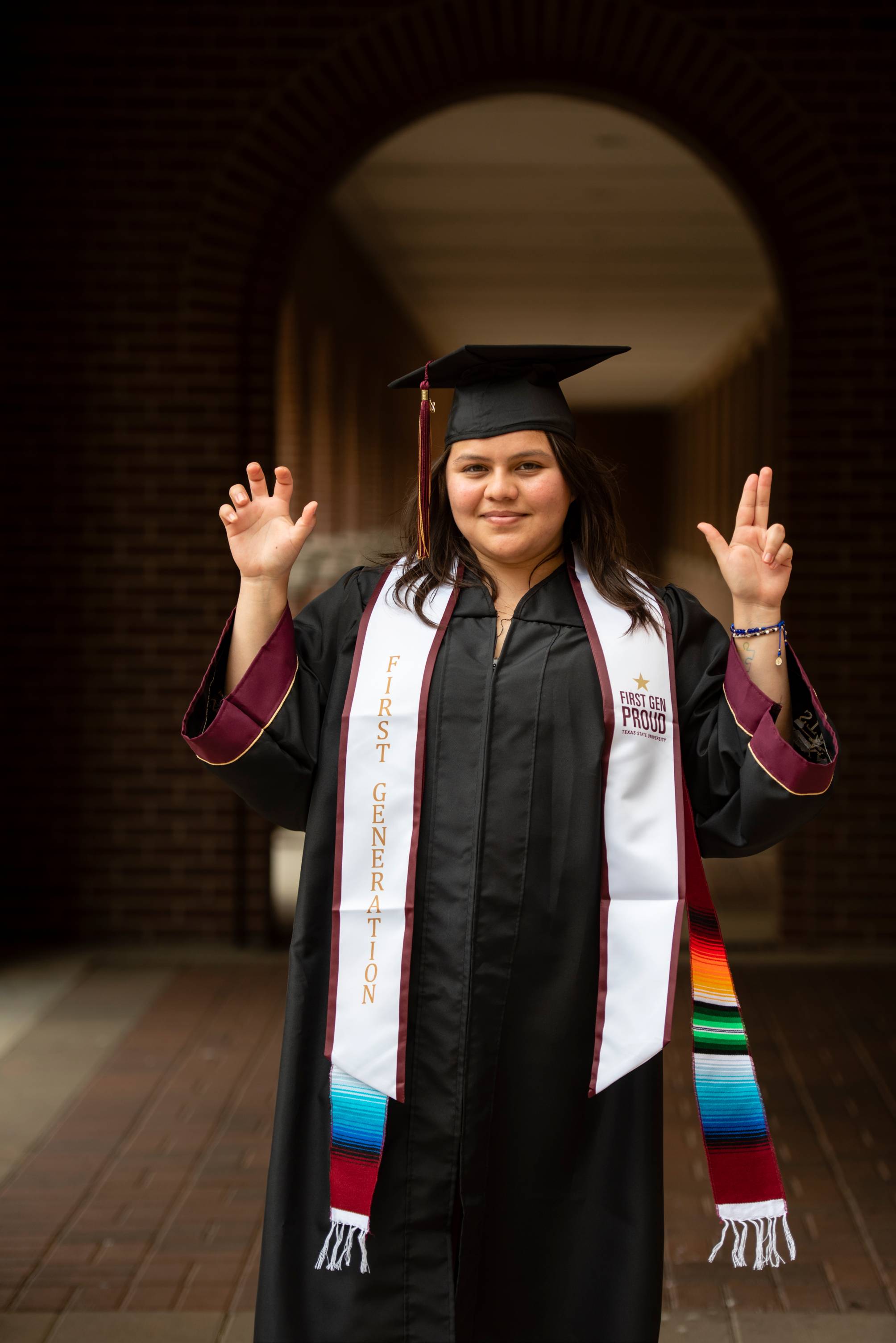 woman in graduation regalia holds up txst hand signs in front of large brick arch