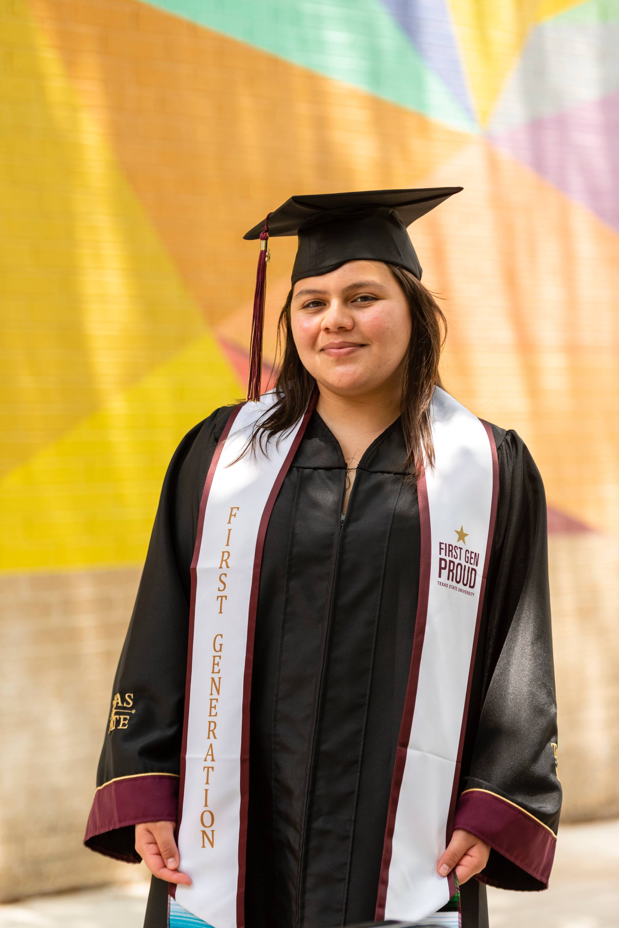 Graduate in cap and gown wearing a First Generation stole smiles at the camera in front of the colorful diamond-patterned One of a Kind mural on campus.