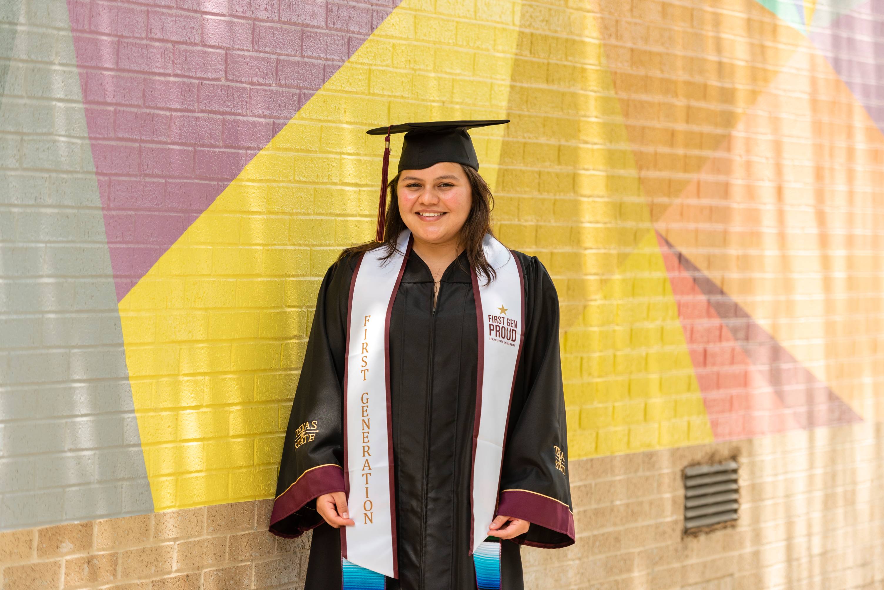 Graduate in cap and gown wearing a First Generation stole smiles at the camera in front of the yellow and geometric-patterned One of a Kind mural.
