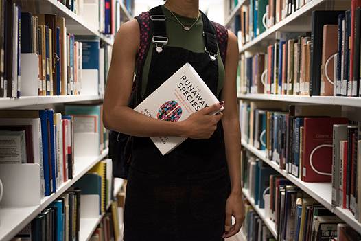 student wearing black overalls holding a textbook between bookshelves in a library