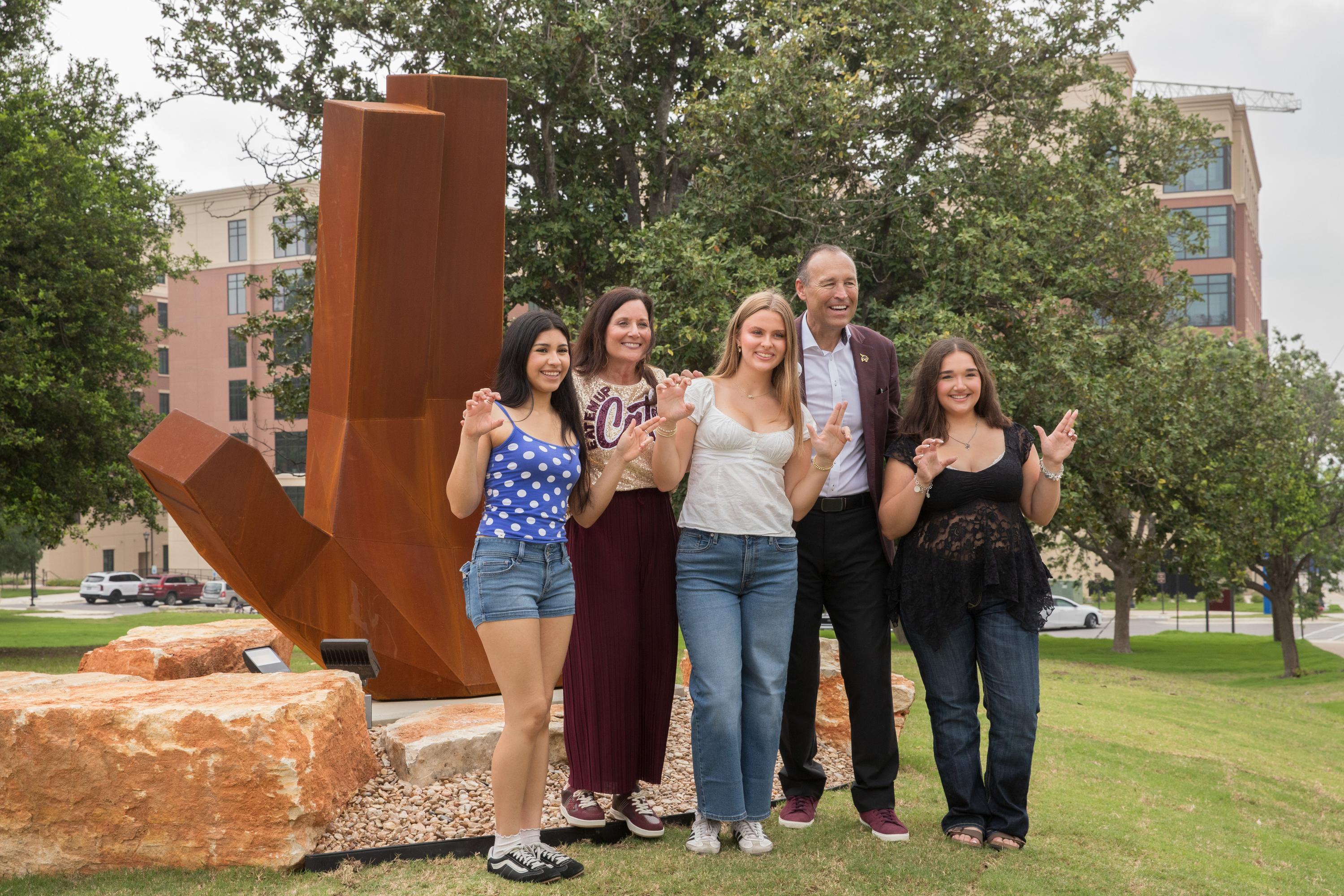 five people posing in front of large steel statue while holding up txst hand signs