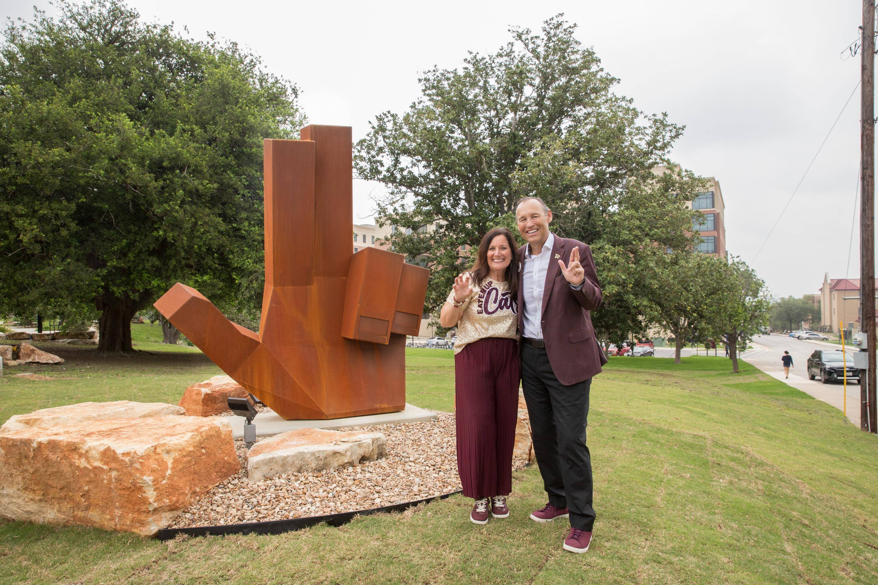 kelly and beth damphousse stand next to large steel statue while holding up txst hand signs