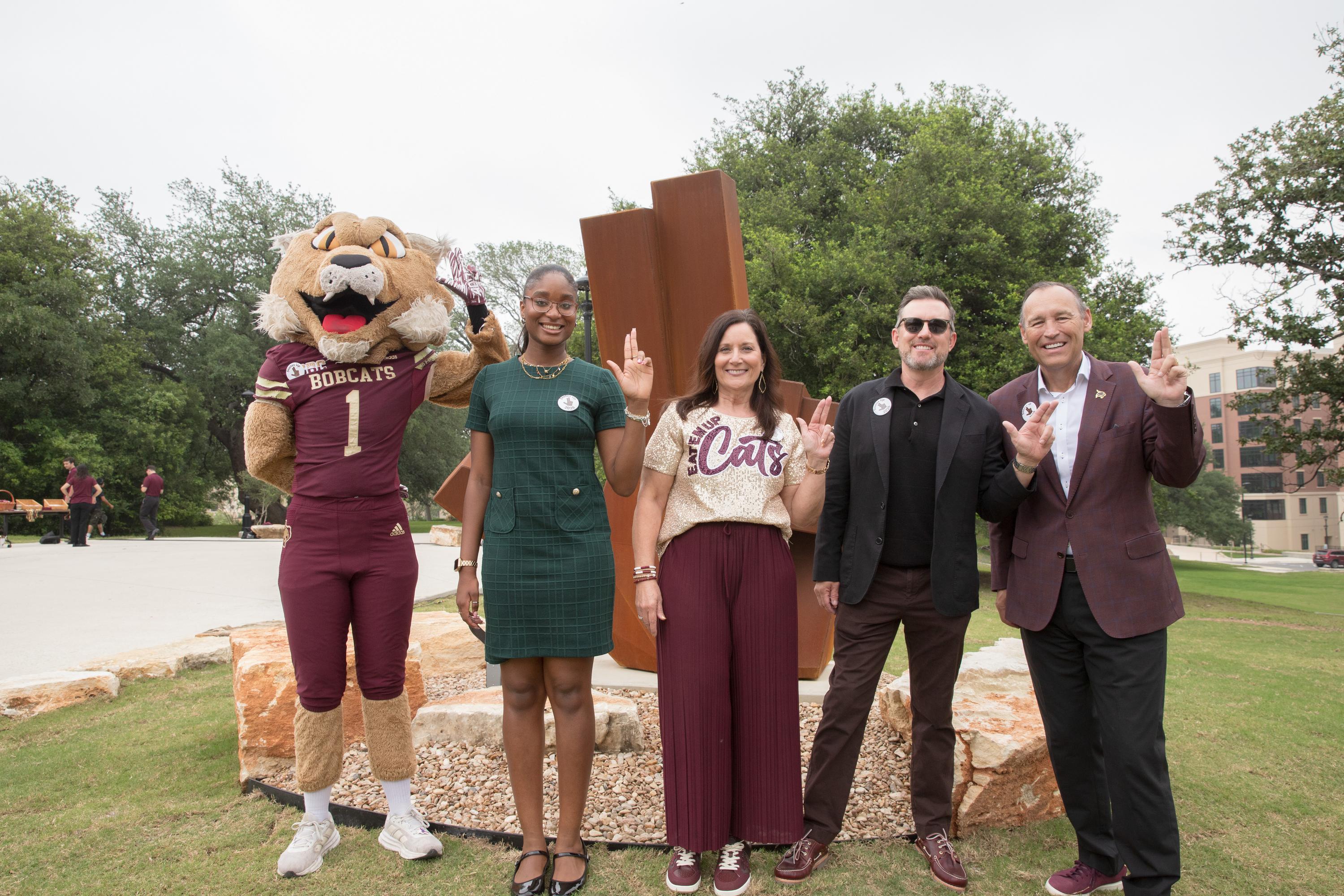 four people and a bobcat mascot stand in front of a large steel sculpture shaped like a hand