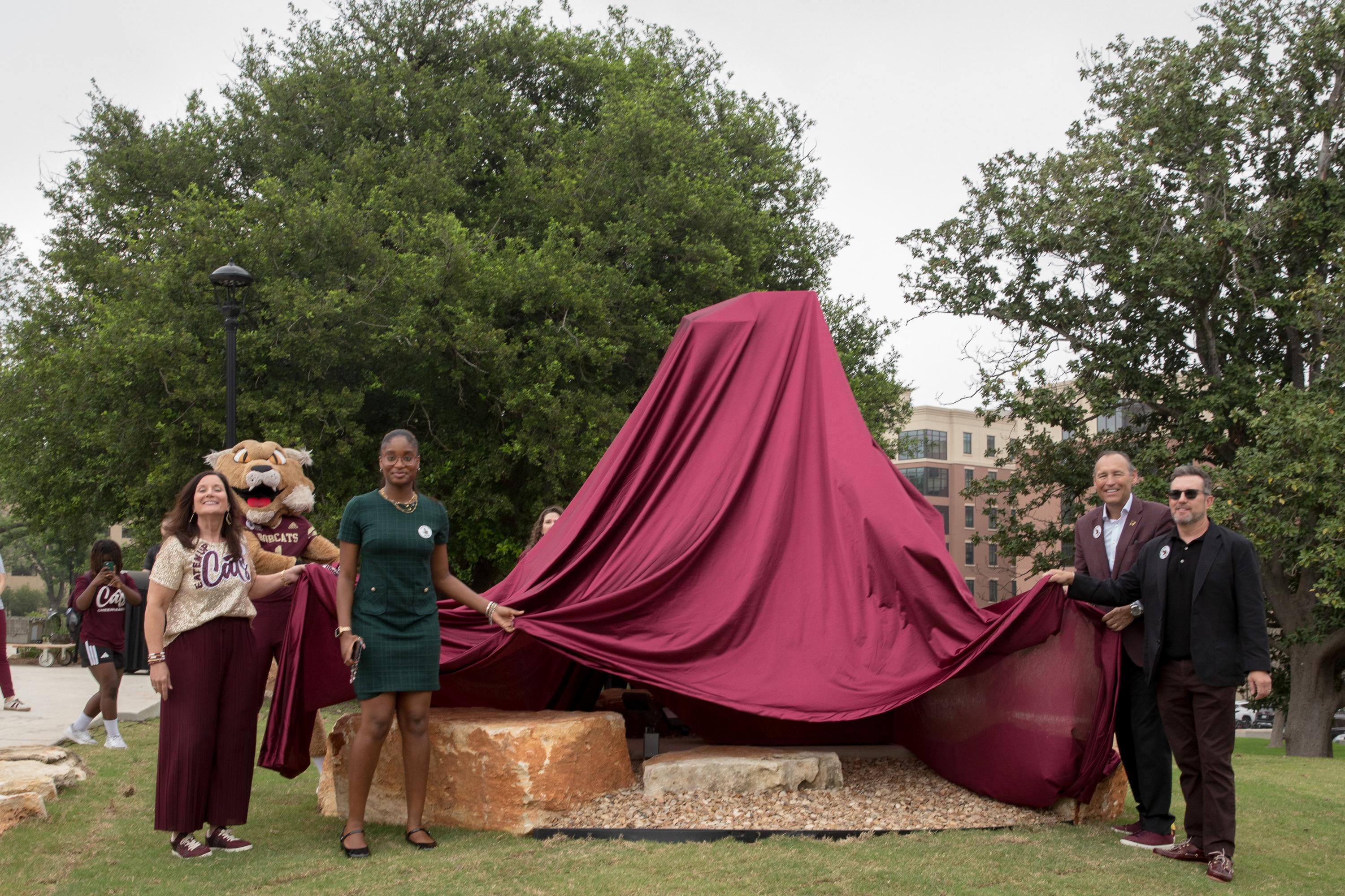 four people and a bobcat mascot stand next to a large sculpture that is covered in a large maroon fabric cover