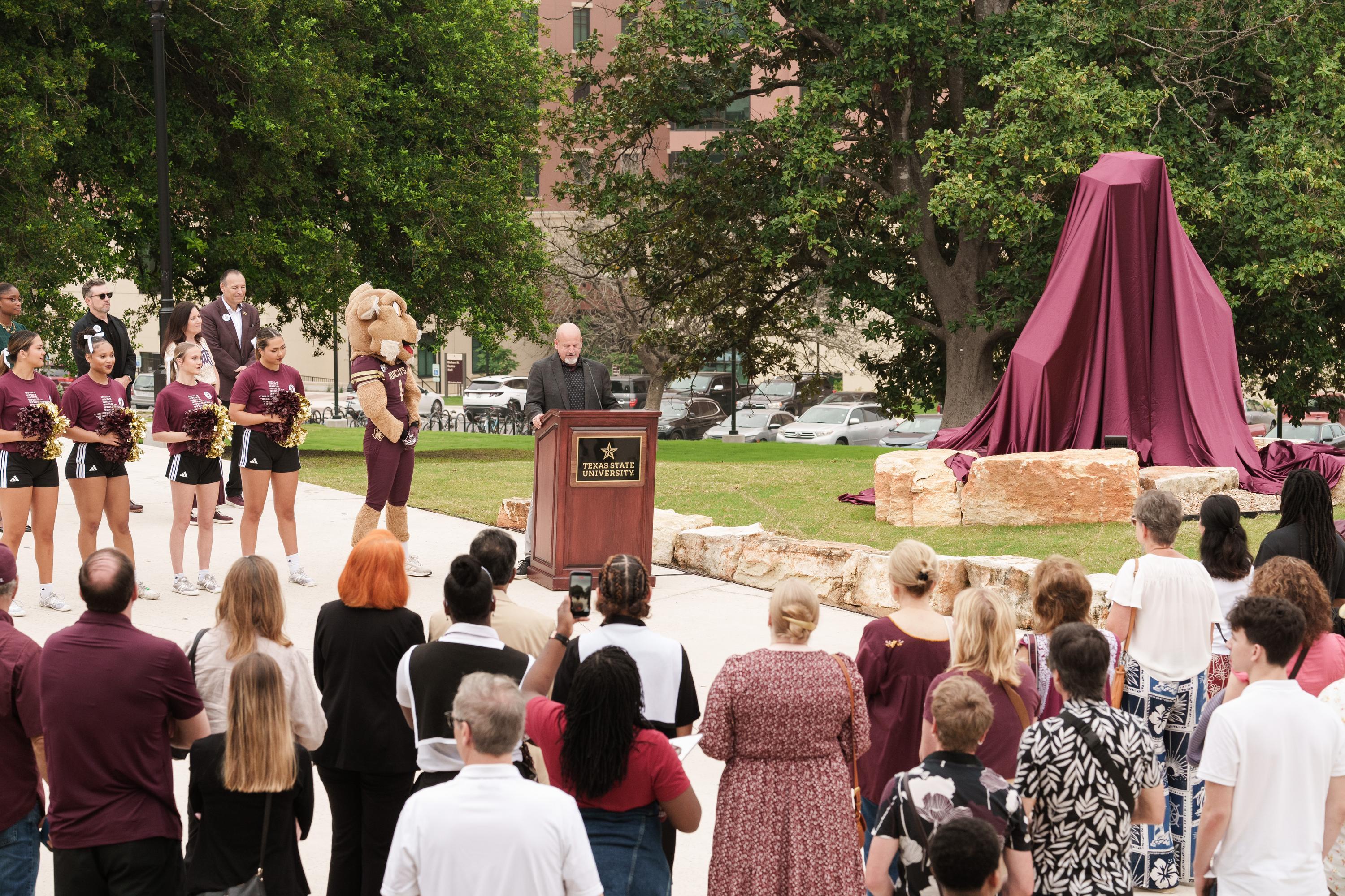 man gives speech behind wooden podium. a large group is gathered around with a larged covered sculpture in the background.
