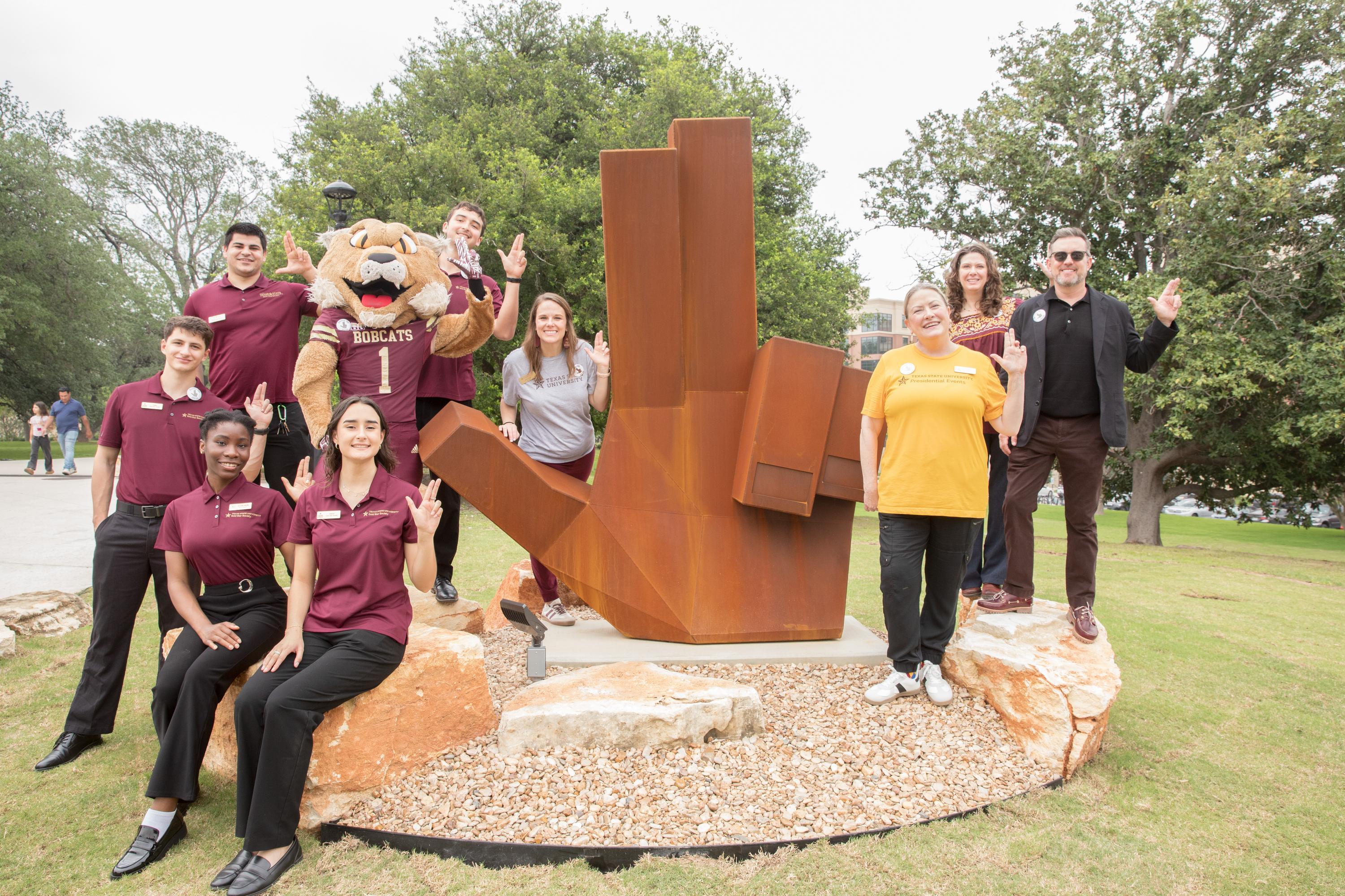 large group of people posing for photo in front of large steel sculpture that is shaped like the txst hand sign.