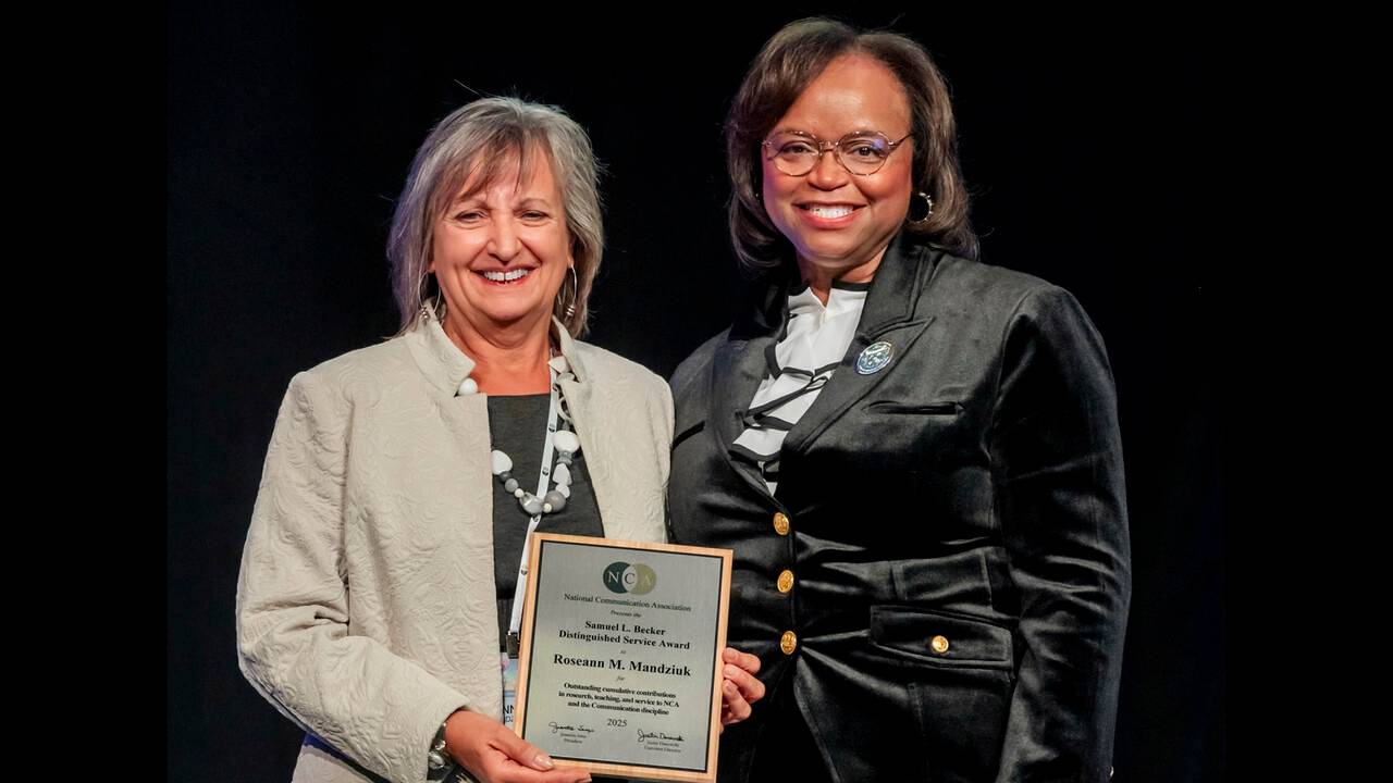 two women smiling and posing in front of black backdrop. women on left is in grey holding an award. the women on the right is in black smiling.
