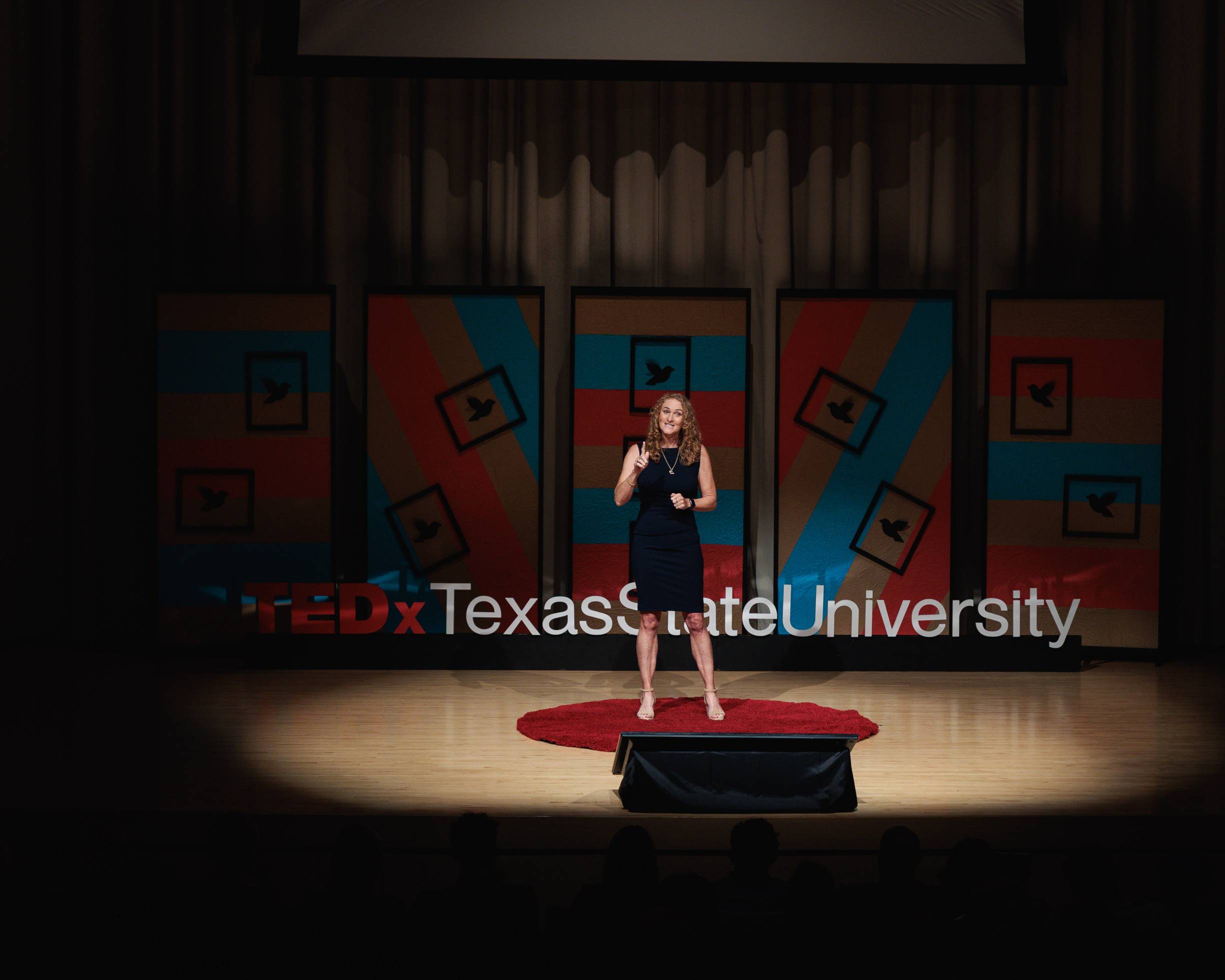 women in black on dim lit stage speaking in front of an audience. The stage had a backdrop of blue and red with the tedx texas state university in large letters at the bottom