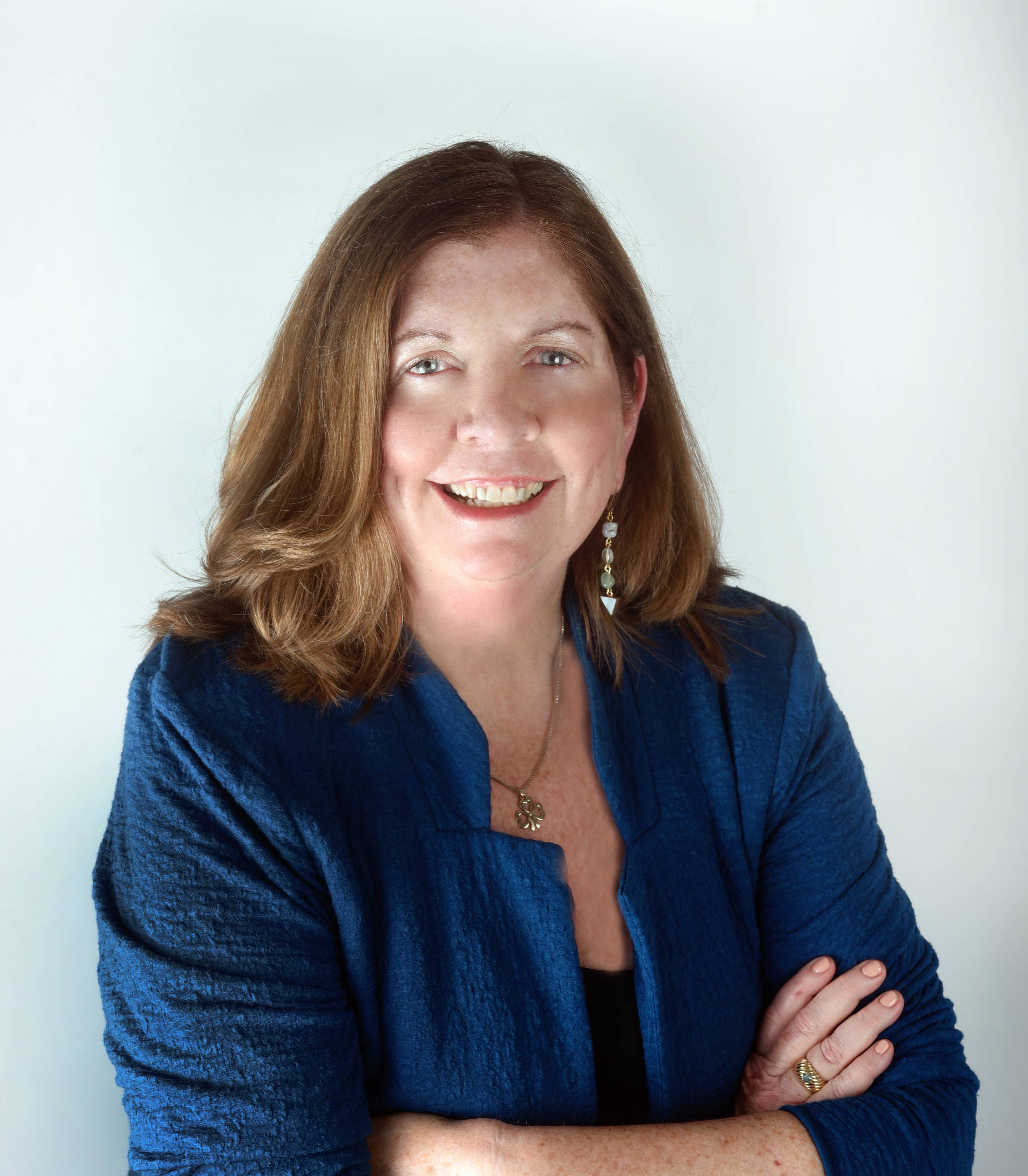 woman smiling in blue blazer and shirt in front of white background with her arms crossed