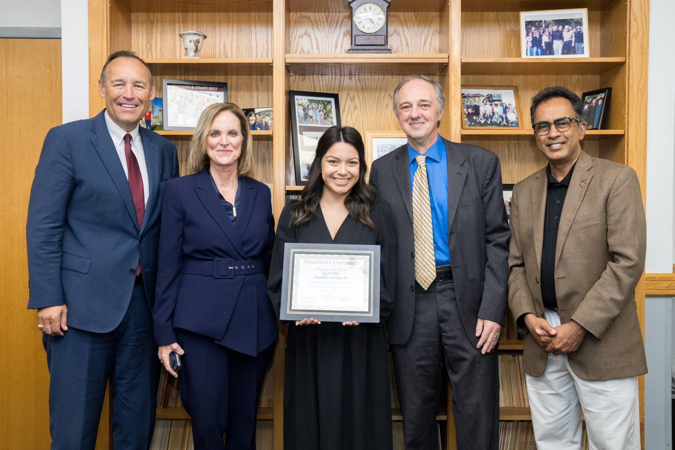 5 people standing and smiling in a line. the person in the middle is holding an award for staff employee of the month