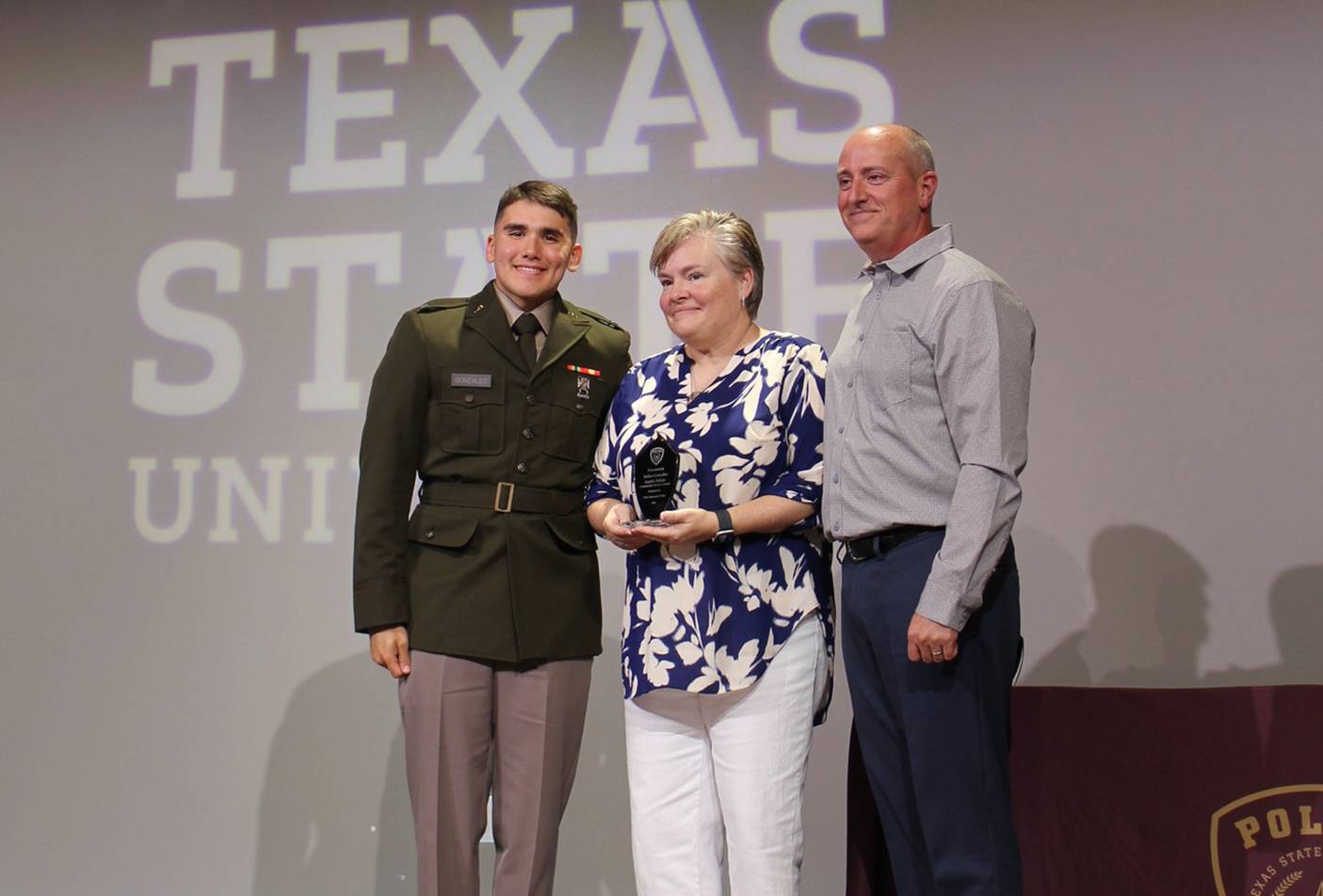 man in military uniform standing on stage with man and woman. the woman, who is standing in the middle, holds up an award