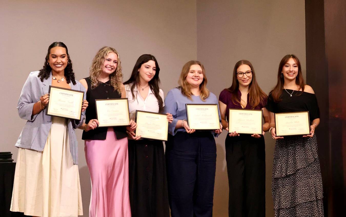 six women hold up certificates on stage
