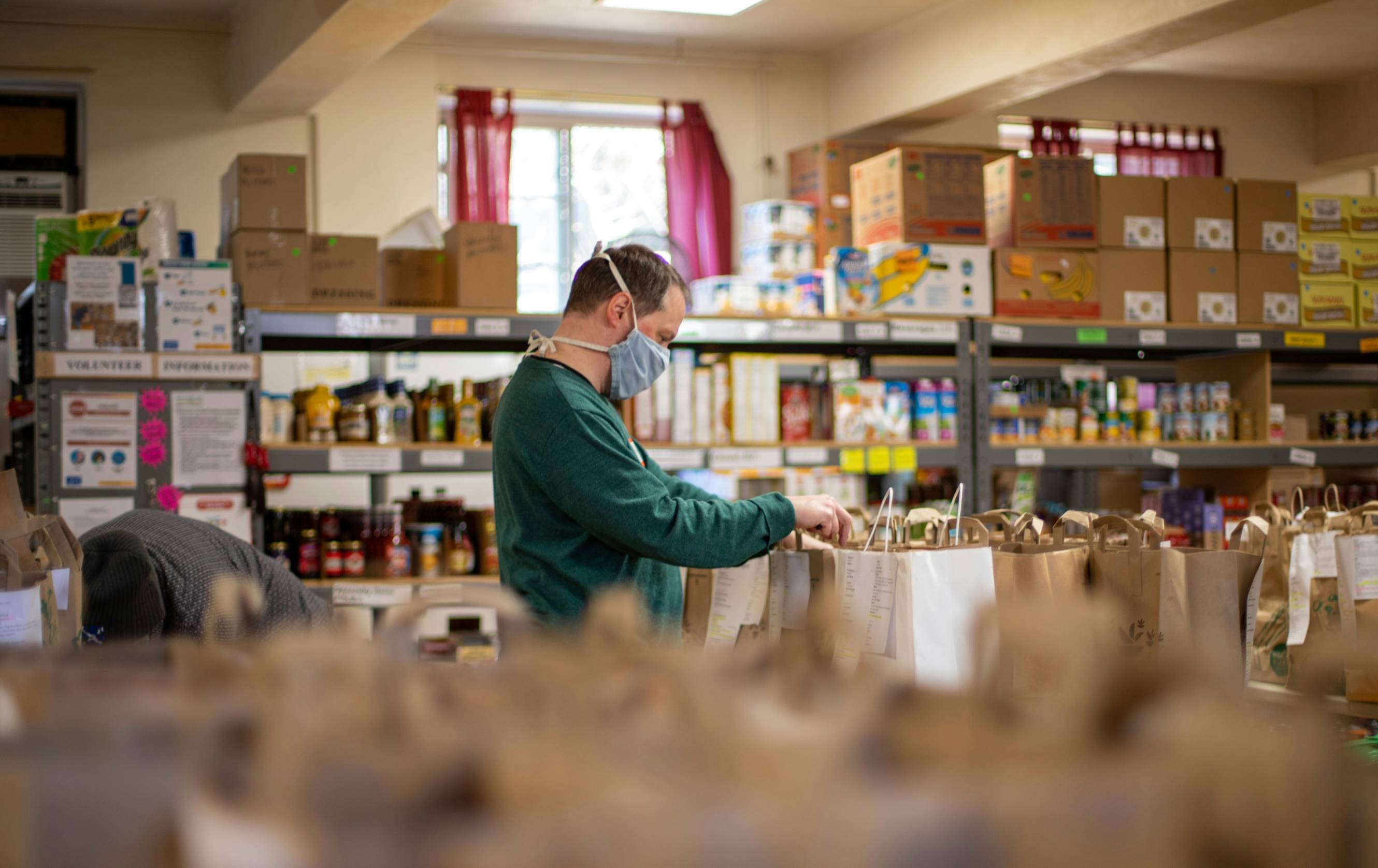 A volunteer wearing a face mask organizes grocery bags inside a community food pantry lined with shelves of donated food and supplies.