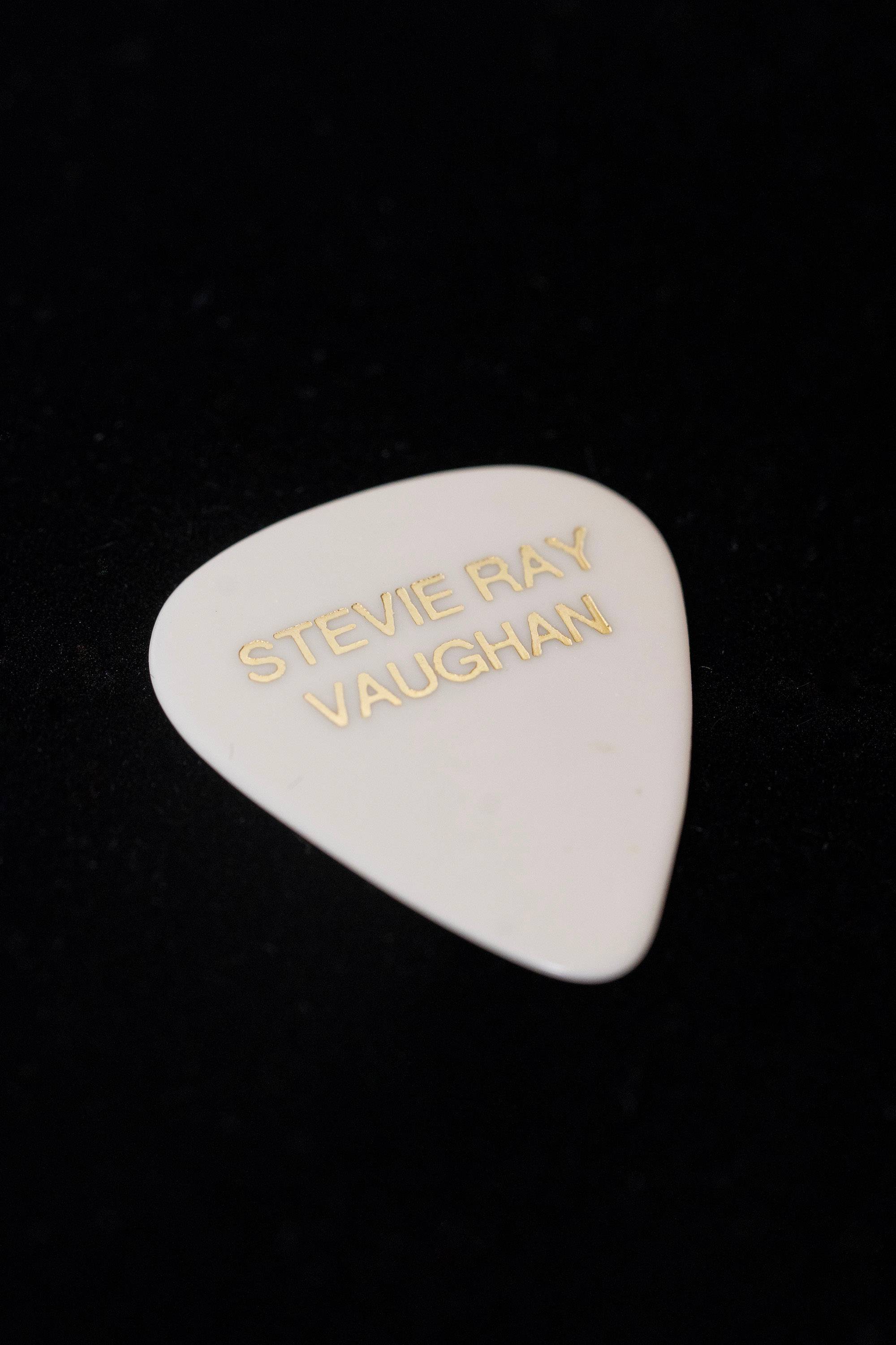 A white guitar pick with the name Stevie Ray Vaughan on it colored in gold.