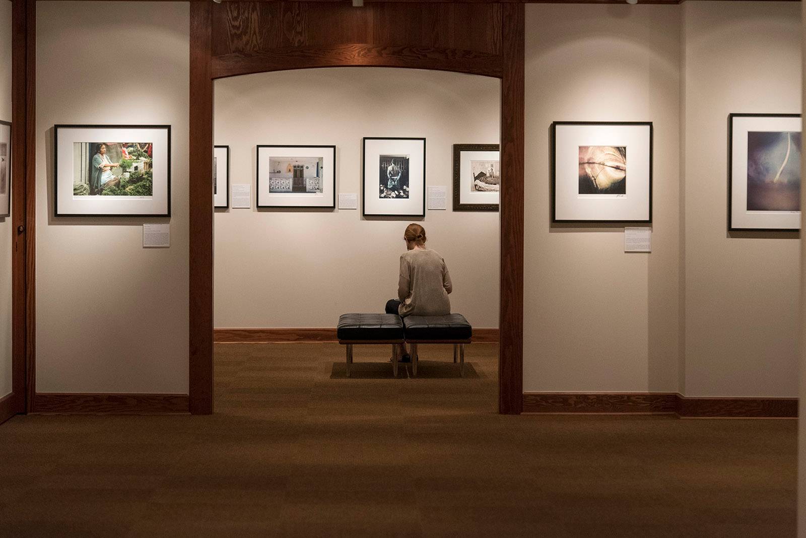 A woman sits on a bench in the middle of a gallery surrounded by photography on the walls.