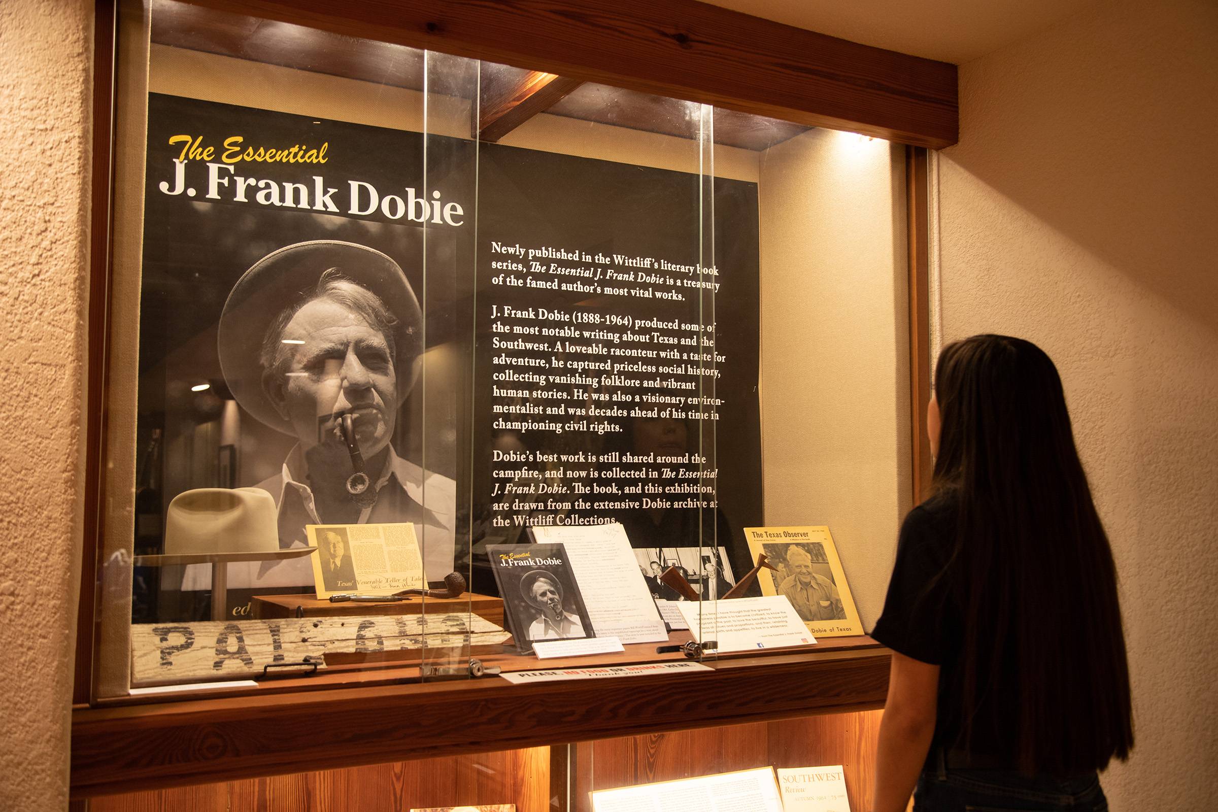 A woman stands in front of a glass case containing a large black and white portrait of J. Frank Dobie and some of his personal artifacts.