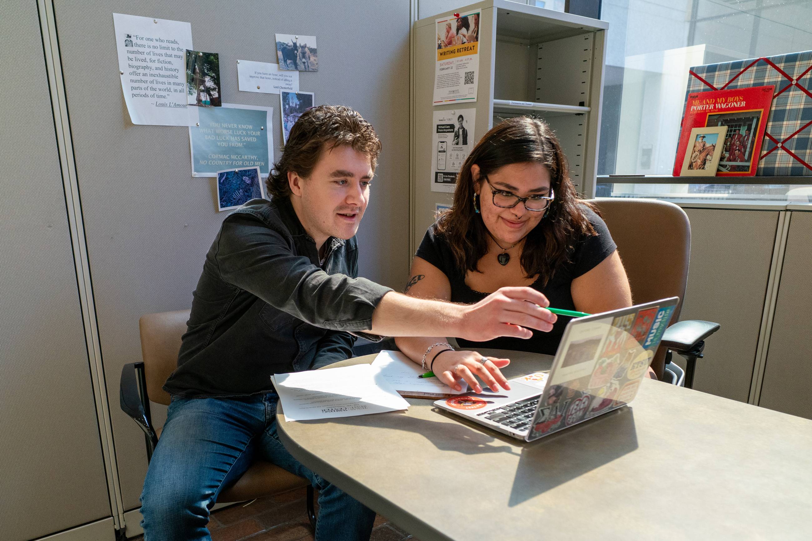 2 people sitting at a desk reviewing a paper on a laptop at the writing center at TXST.