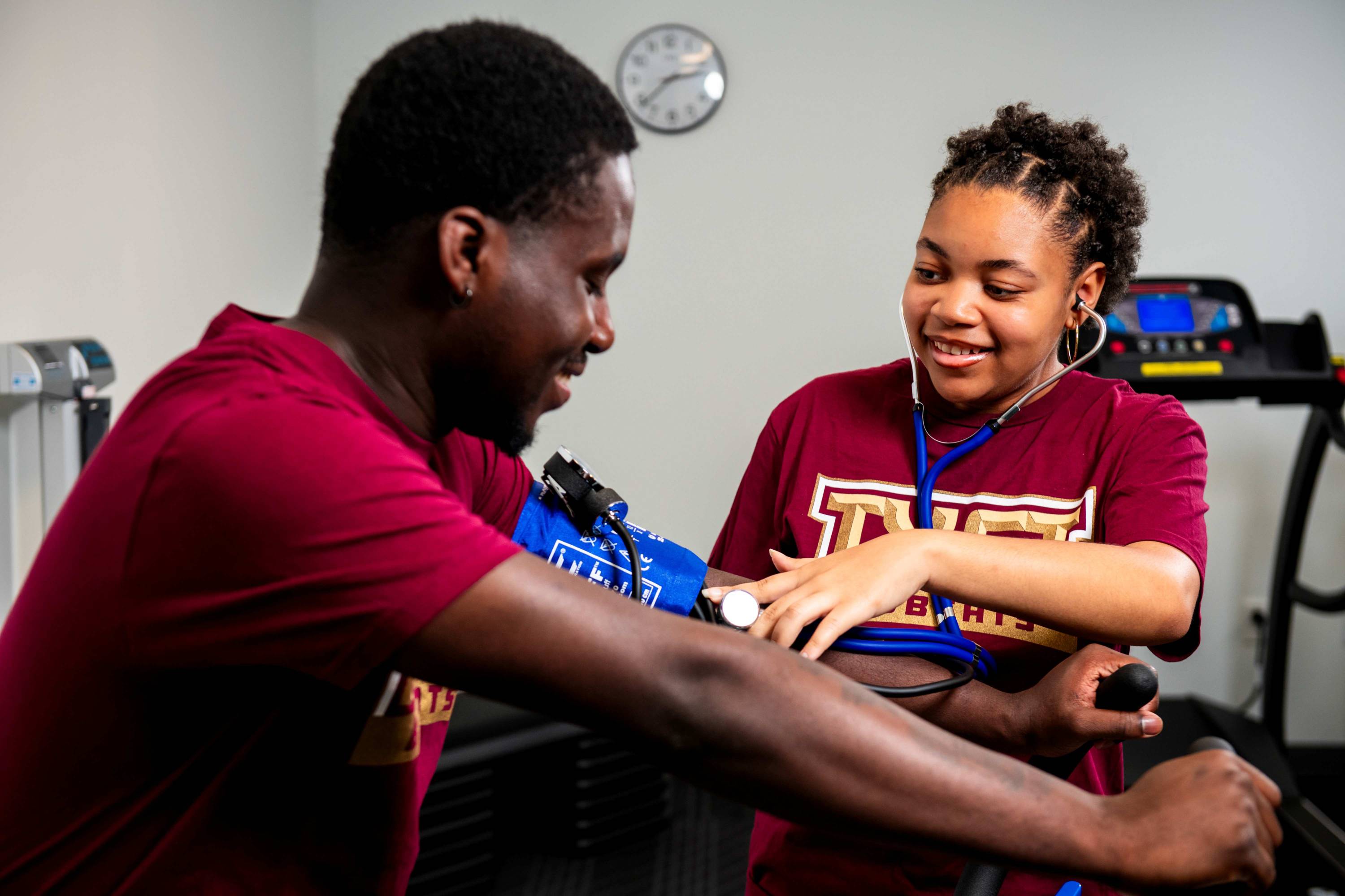a woman wearing a stethoscope and a maroon TXST branded tee shirt measures the blood pressure of a man using a blue arm cuff