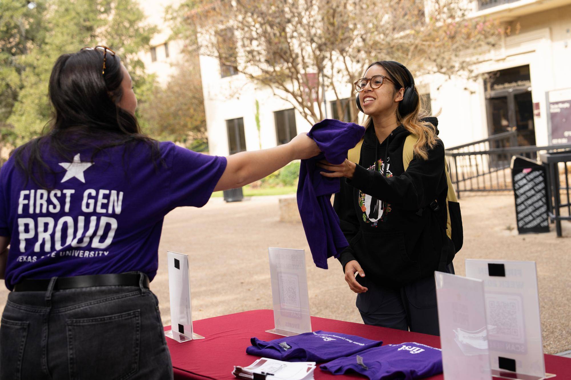 a student wearing a t-shirt reading "first gen proud" on the back hands another student that same shirt at an outdoor tabling event.