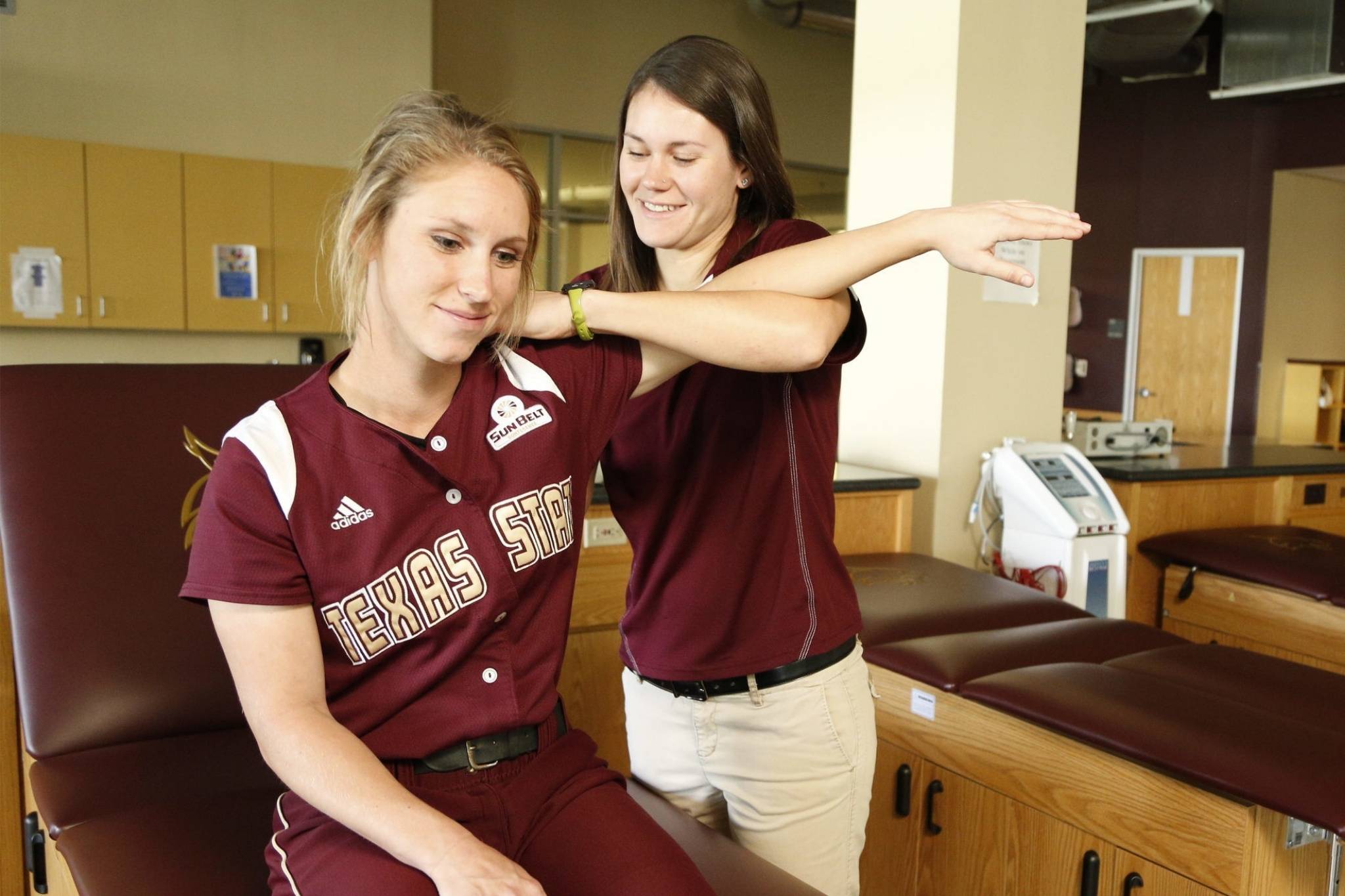 A woman in a softball outfit sitting on an exam table has her shoulder stretched by another woman in a maroon collared shirt in a medical office.