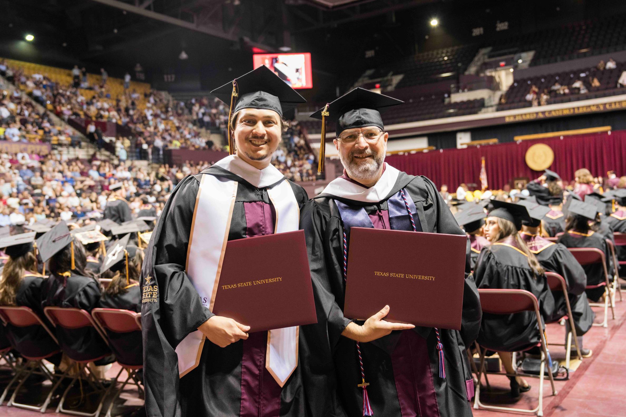 jeff and tyler stand in at commencement in their cap and gown attire holding diplomas