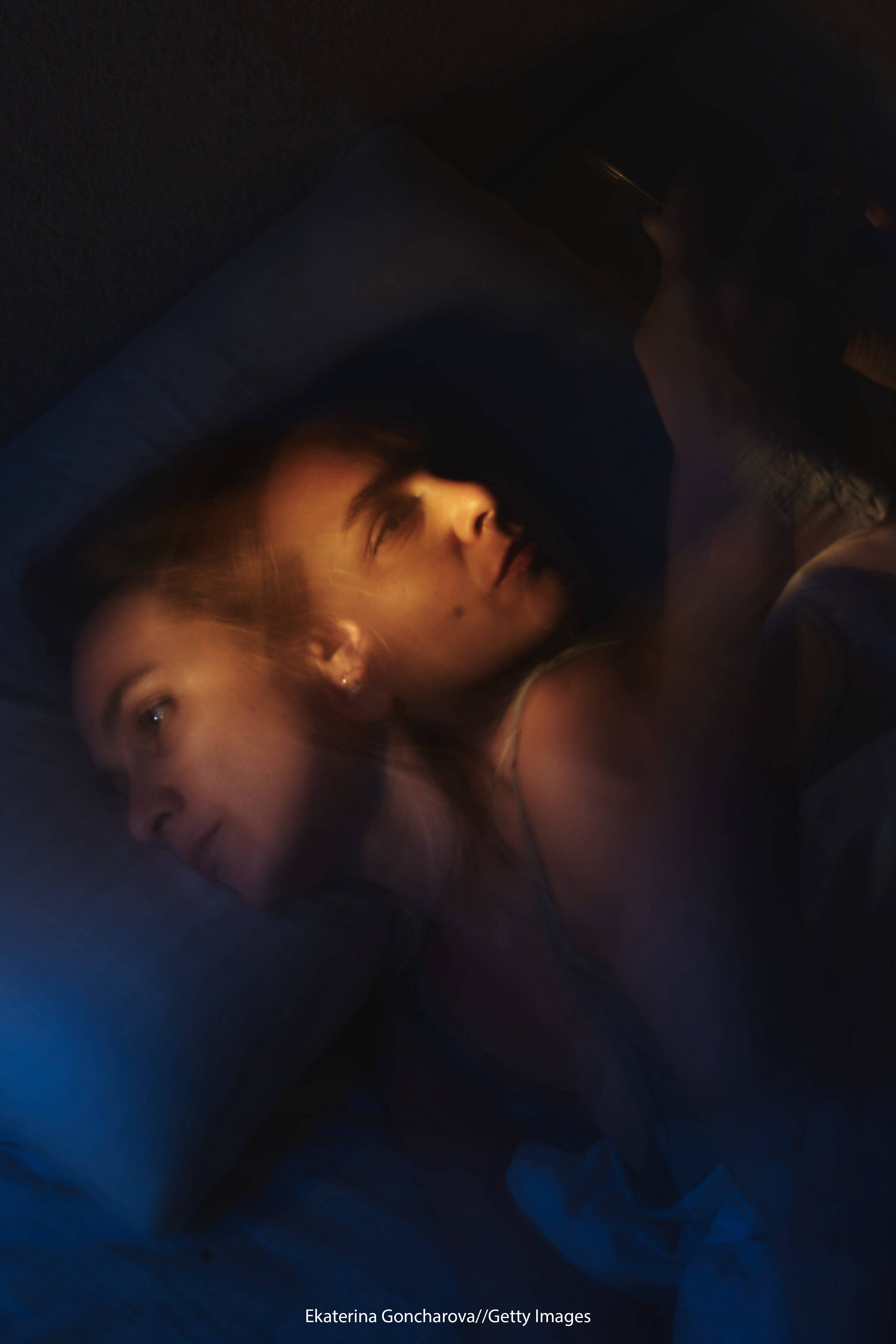 Woman lying awake in bed at night while looking at her phone, with a double-exposure effect showing stress, fatigue, or insomnia.