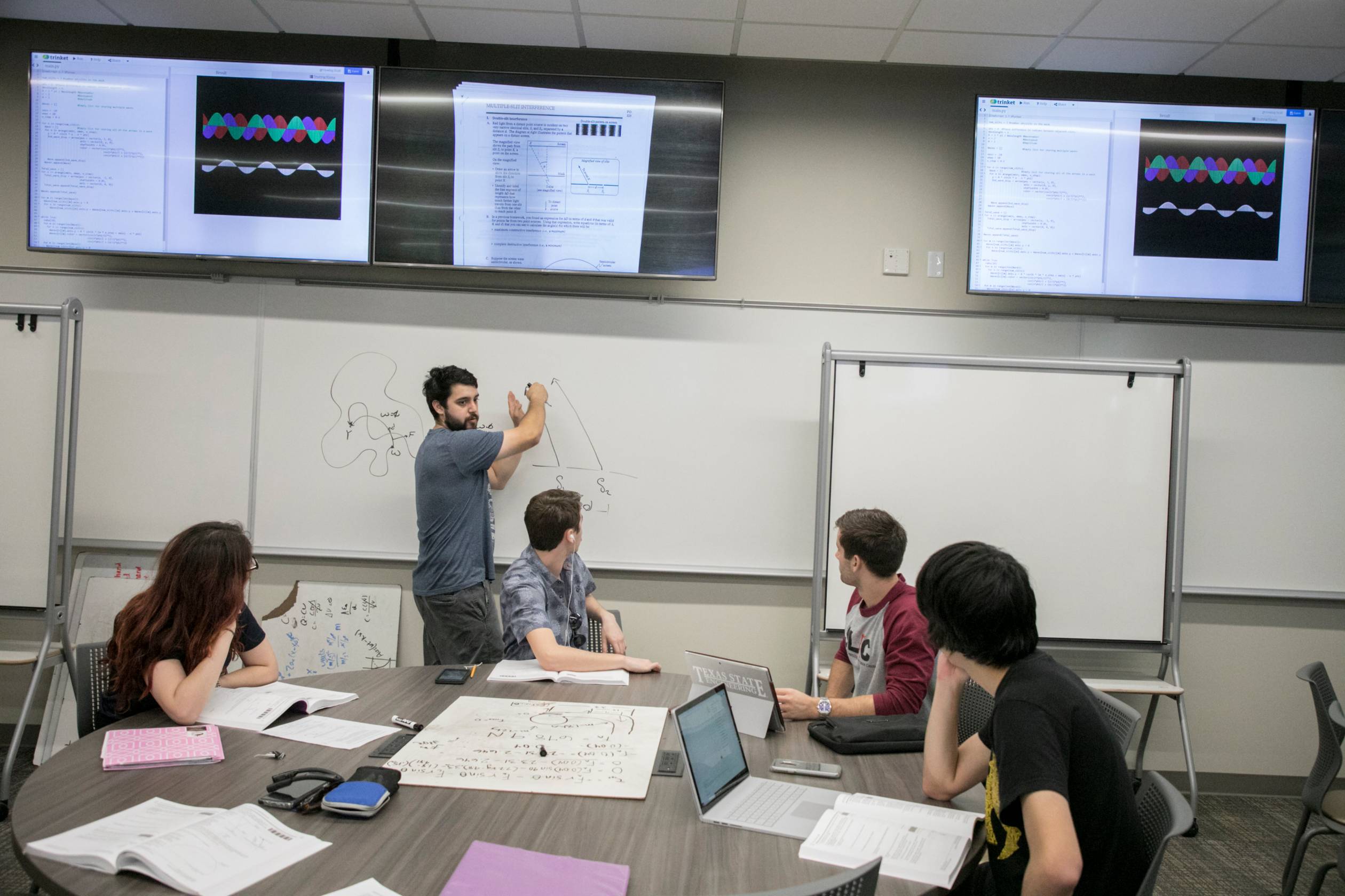 Students at a table listen to another student speaking from graph drawings at a white board.