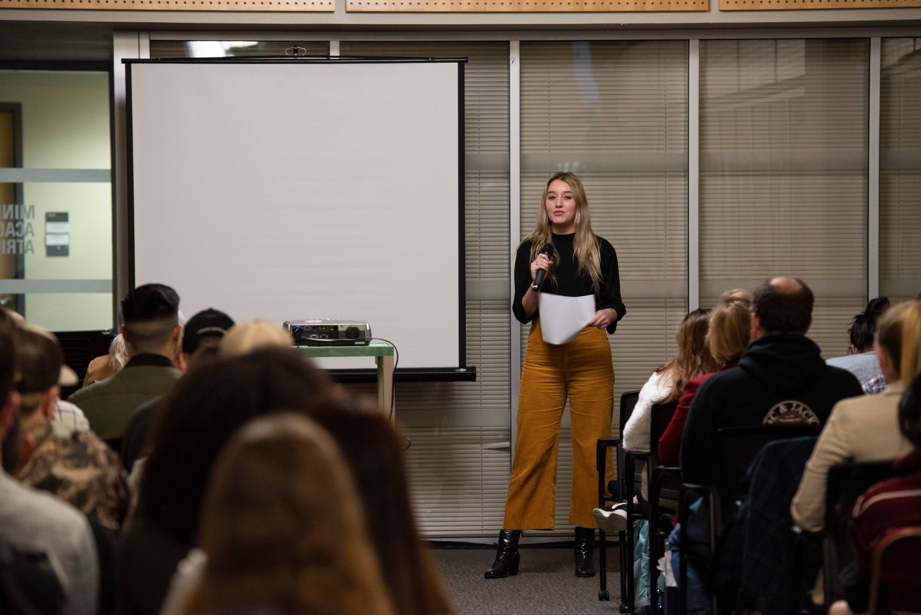 a speaker with a microphone and paper standing in front of a room of seated participants