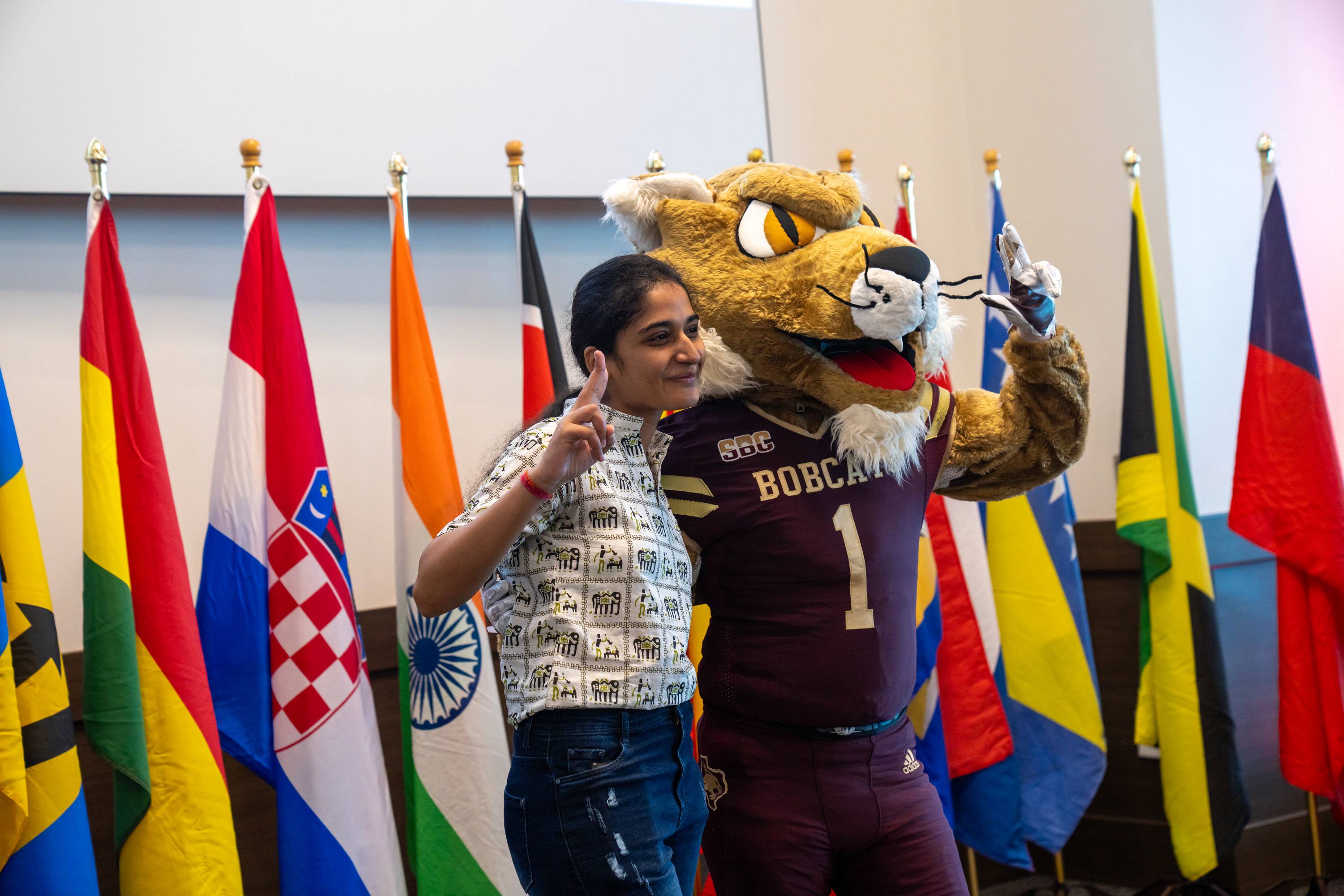 woman posing with bobcat mascot in front of large row of international flags