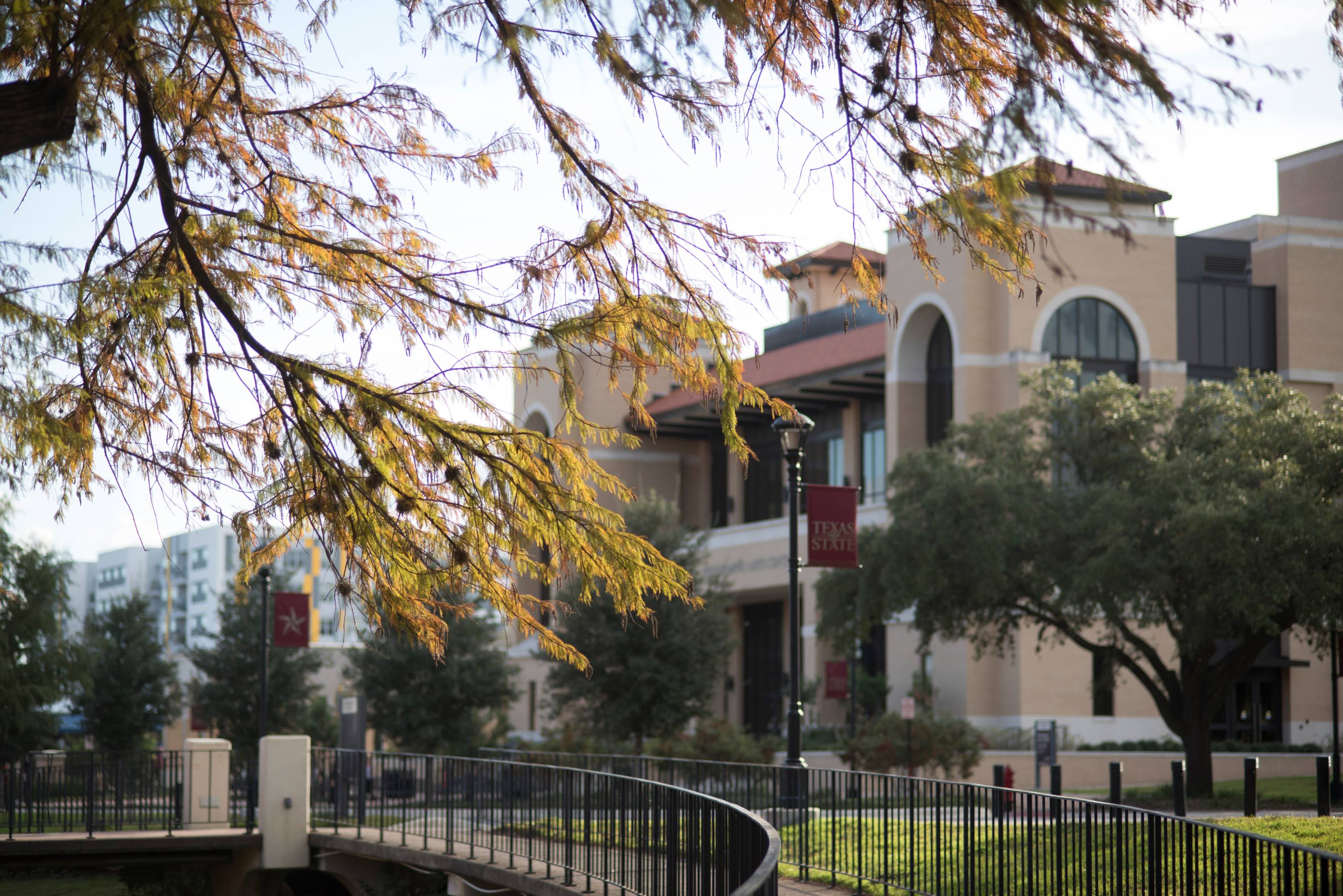 Exterior view of the multi-story Texas State University Performing Arts Center building featuring brick and stucco accents, arched windows, and a paved courtyard walkway. Large shade trees and a black metal fence line the plaza under a clear sky.