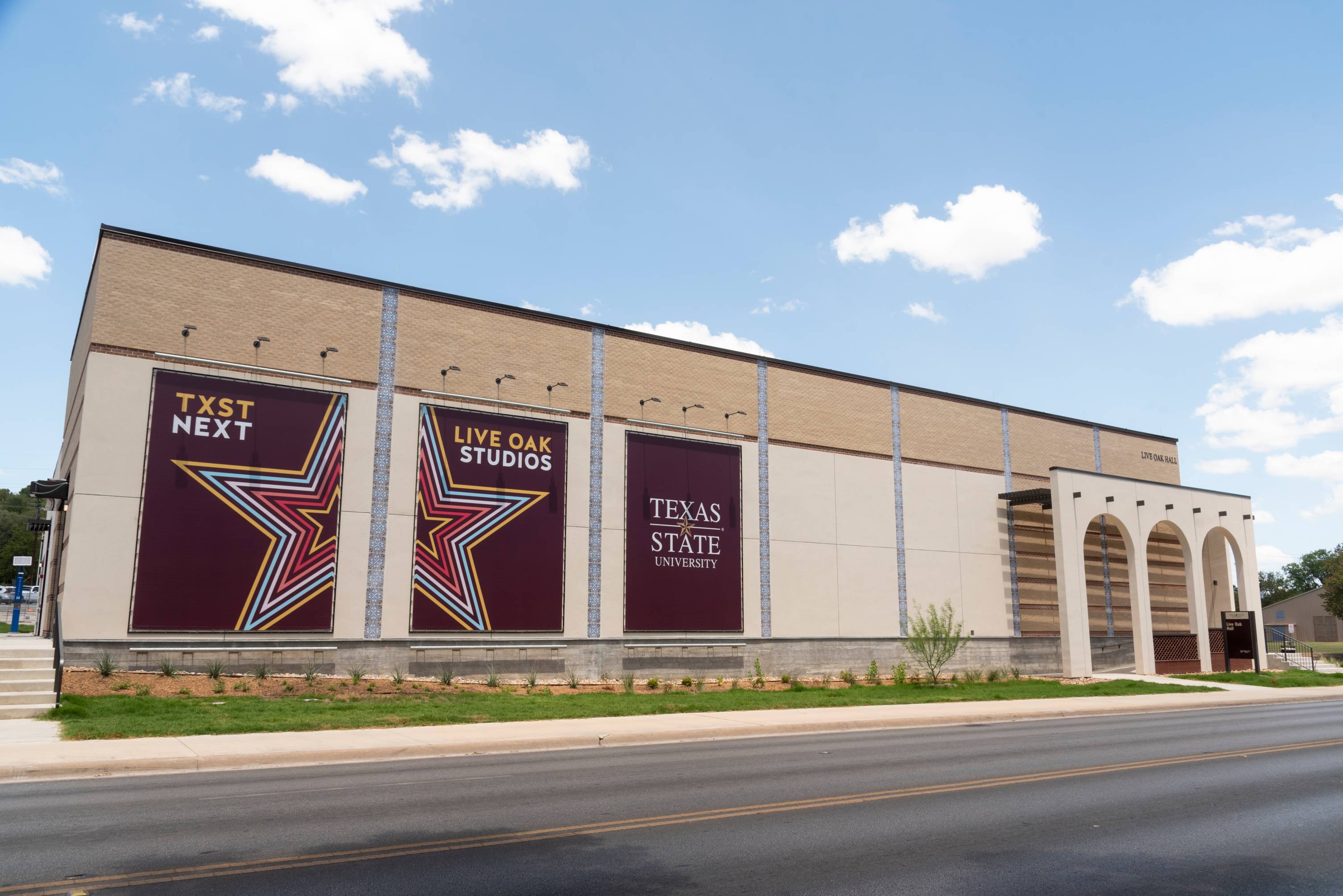 Exterior view of Live Oak Hall at Texas State University, showing large promotional banners for Live Oak Studios on the building's facade. A roadway runs in front of the facility, and a series of arched openings line the right side under a partly cloudy sky.