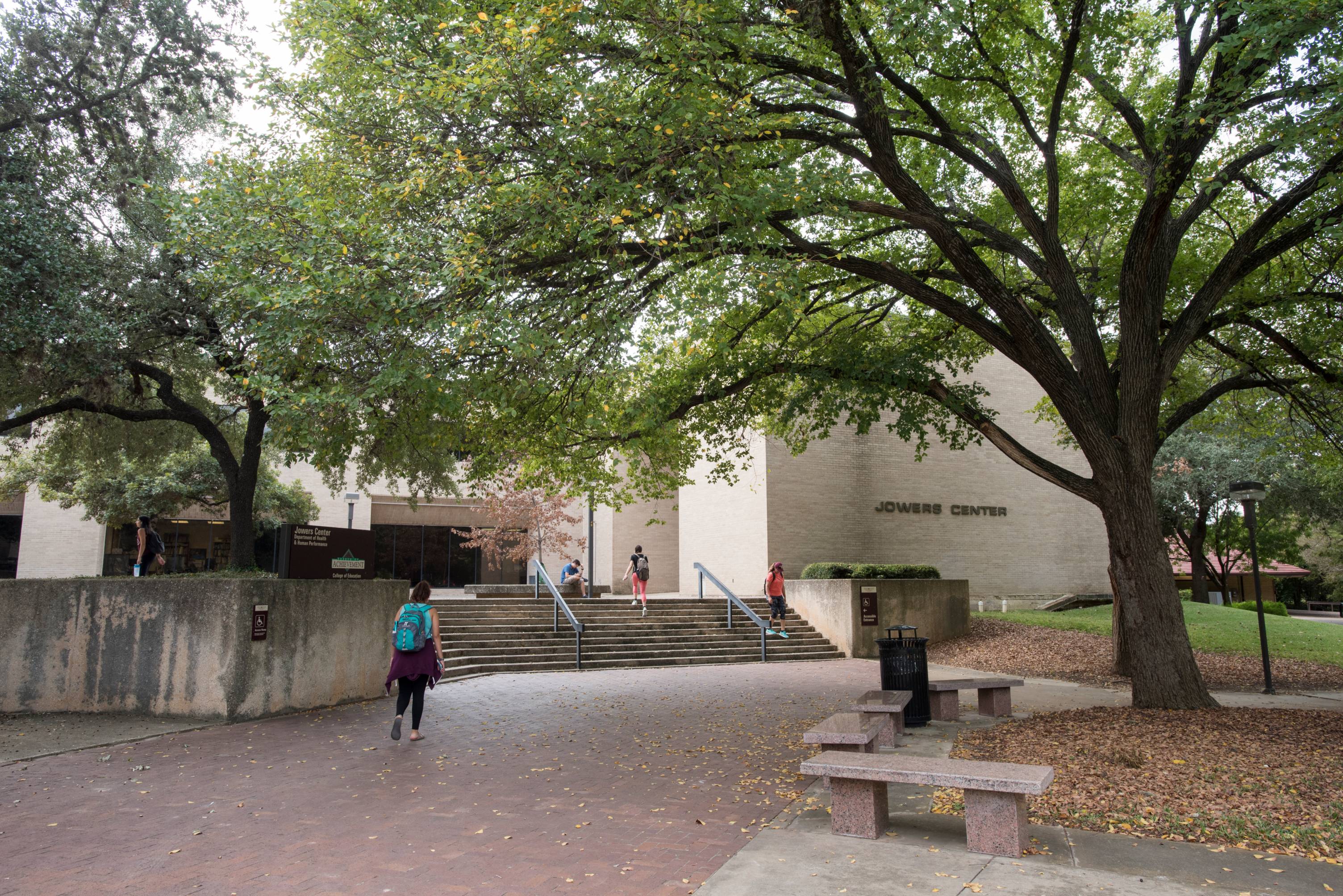 Shaded campus courtyard with large live oak trees and brick paving leading to a wide set of concrete steps. A person with a backpack walks toward the steps ascending to the Texas State University Jowers Center building.