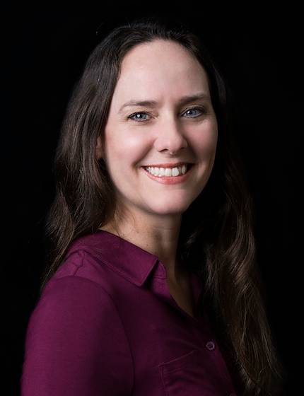 Studio headshot of Michelle Nance smiling, with long dark brown hair and wearing a plum-colored button-down shirt against a dark background.
