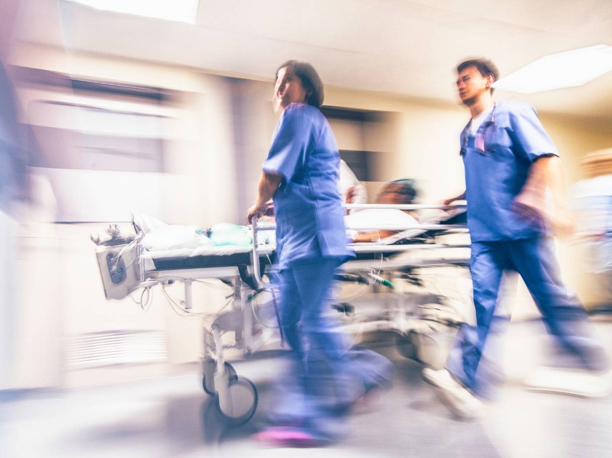 Motion-blurred hospital staff in blue scrubs rush a patient on a stretcher through a brightly lit emergency room hallway, conveying urgency and fast-paced medical care.