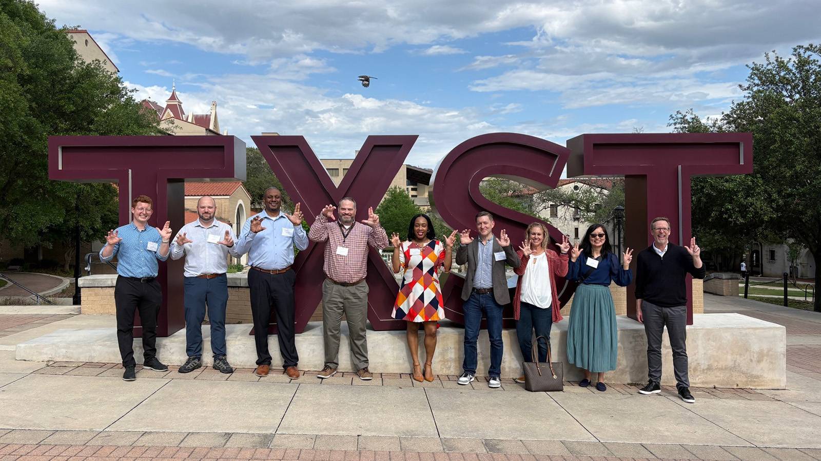 Nine individuals posing outdoors at a large sign that reads "TXST"