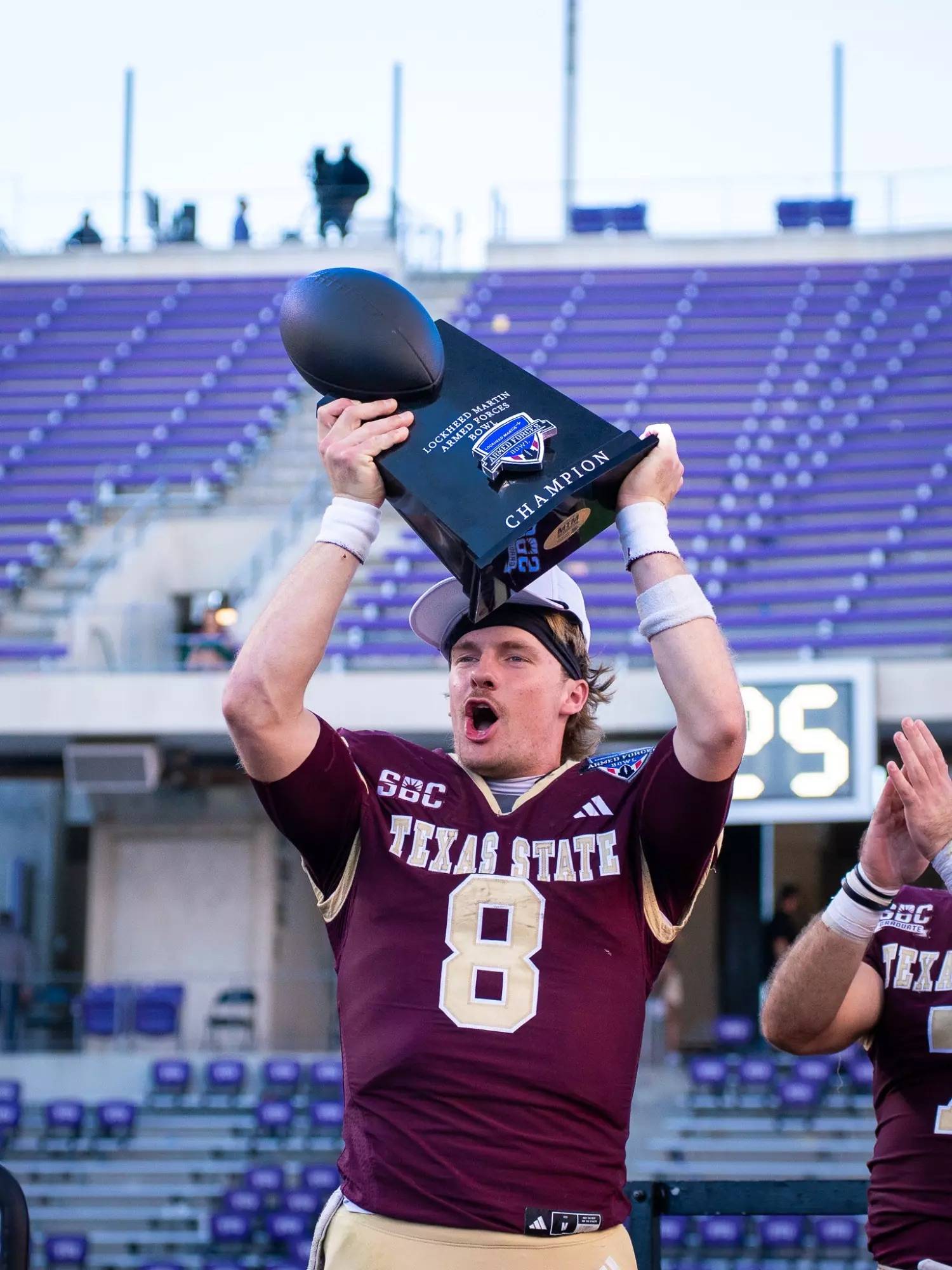 Man in football uniform holds a trophy aloft while football players in maroon uniforms celebrate behind him.