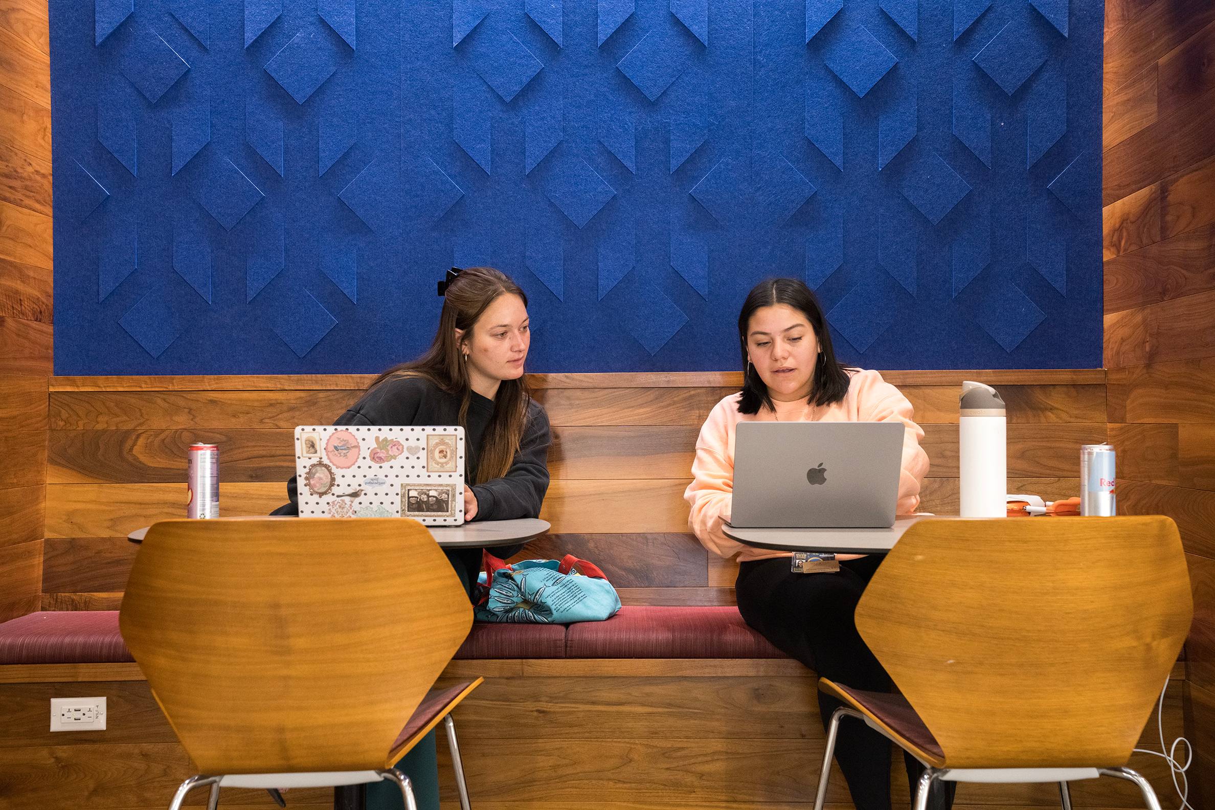 Two students looking at a laptop in front of a blue wall. 