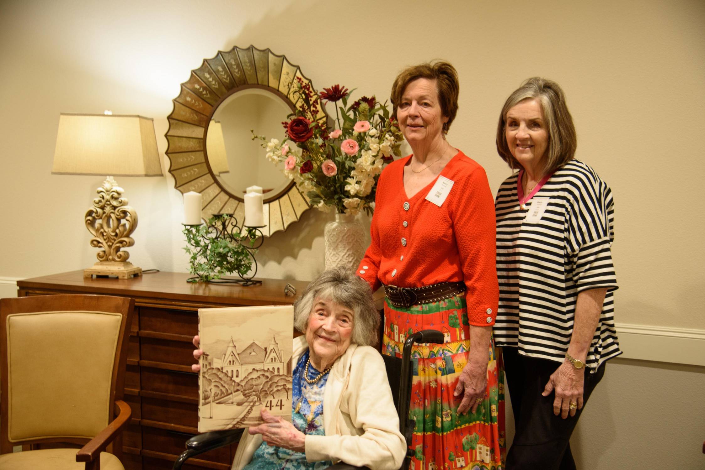 elderly woman in wheel chair holds up a yearbook from 1944 while two women stand behind her smiling