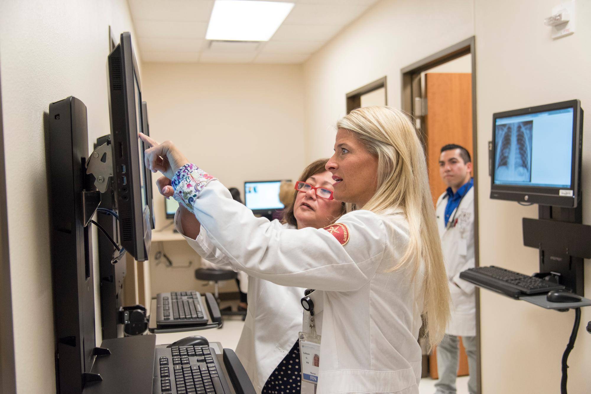 three people in white lab coats examine information on a screen while pointing at it