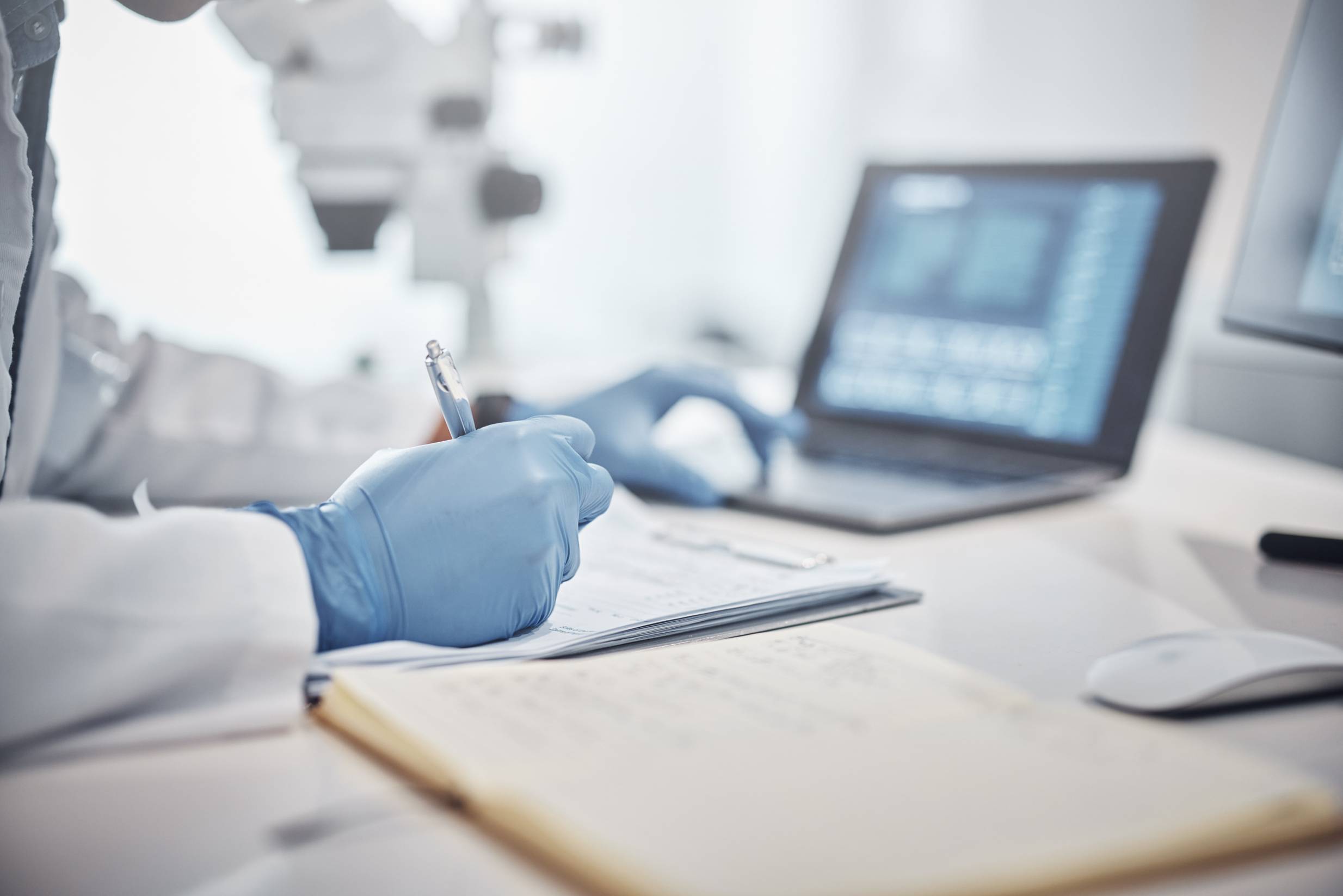 a person wearing a lab coat and blue exam gloves writes in a notebook and uses a laptop computer in a lab environment