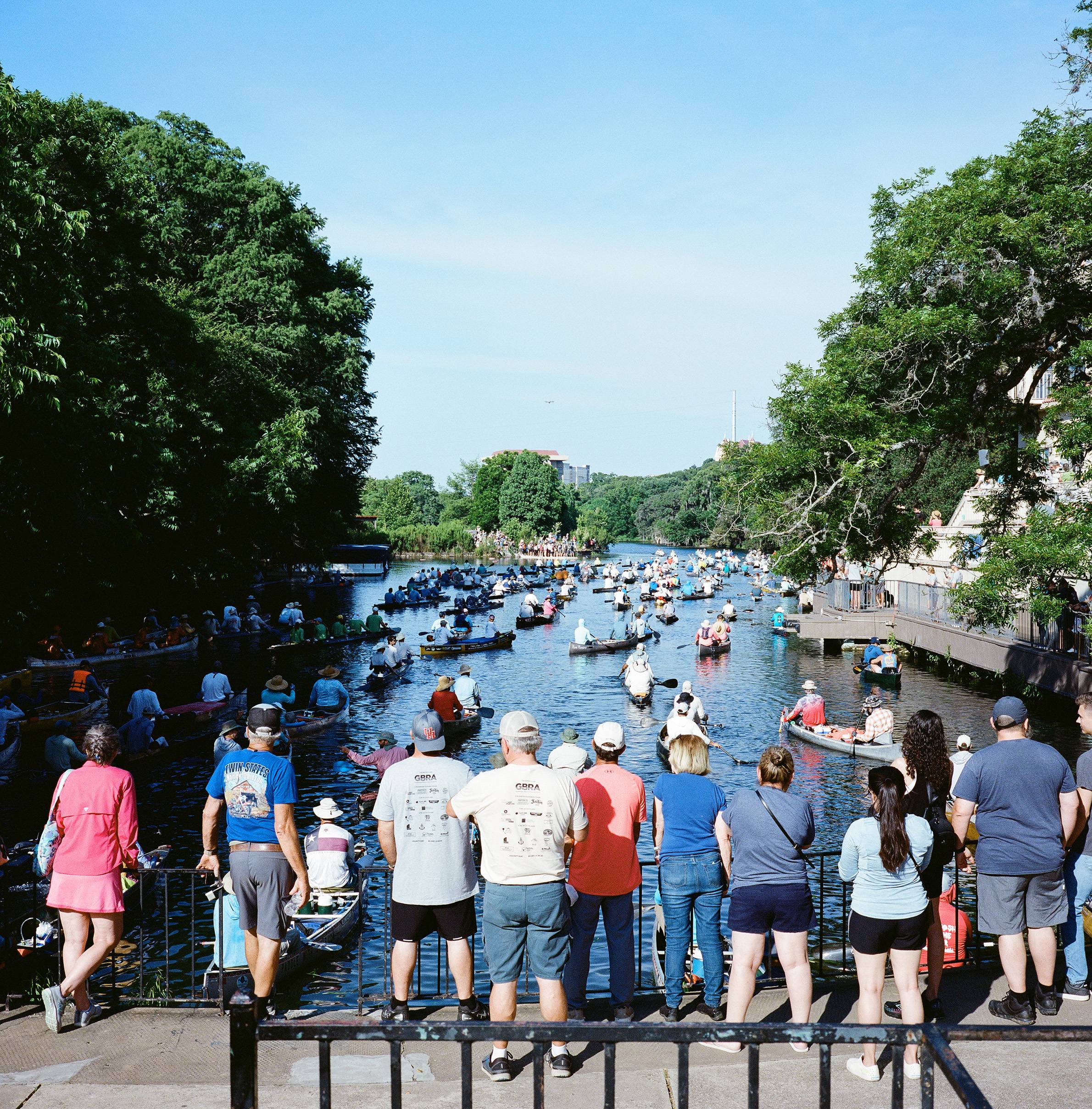 large group of people gather to watch dozens of canoes on san marcos river