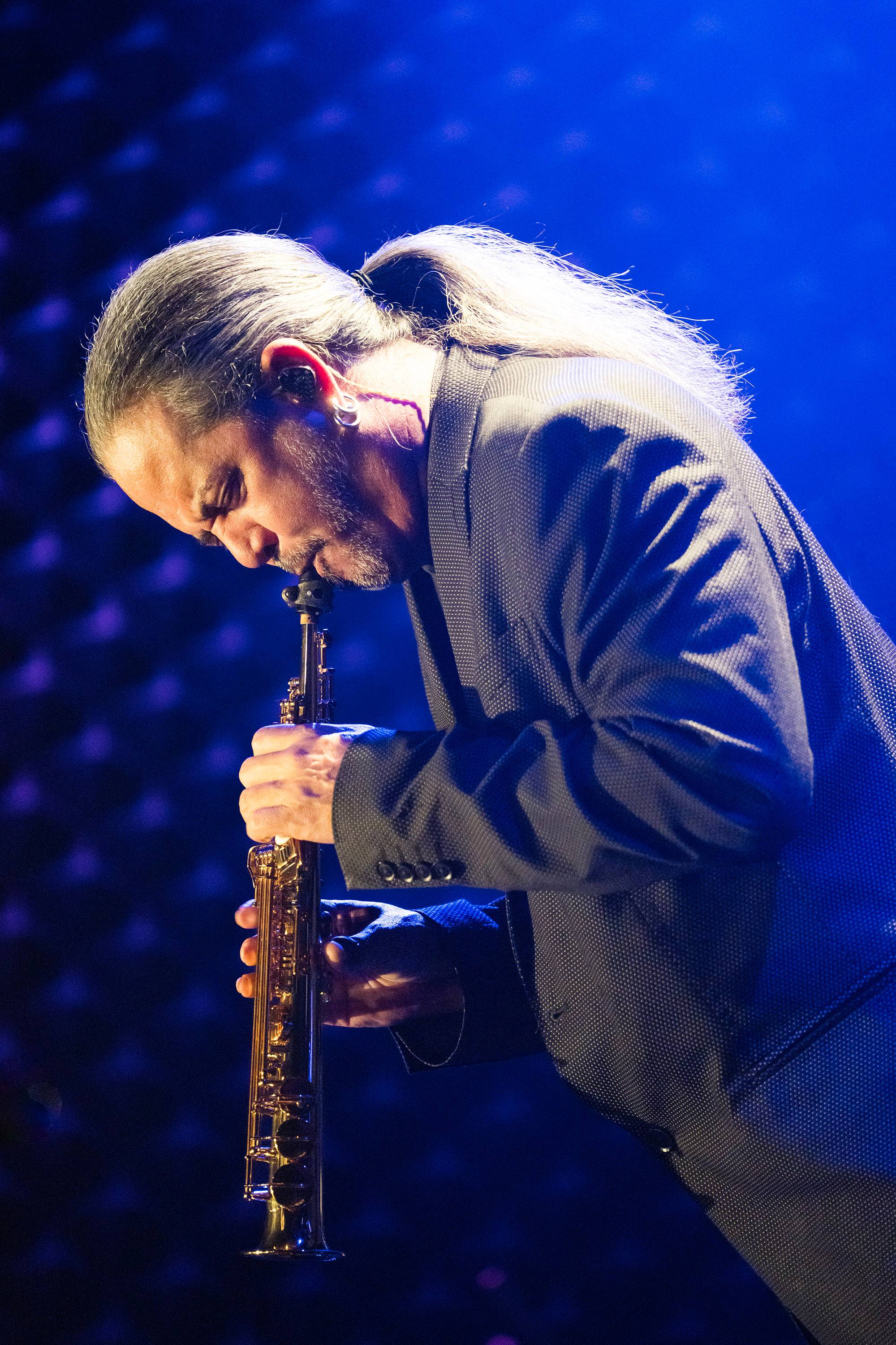 Man with a ponytail is bathed in a blue light and playing the clarinet. 