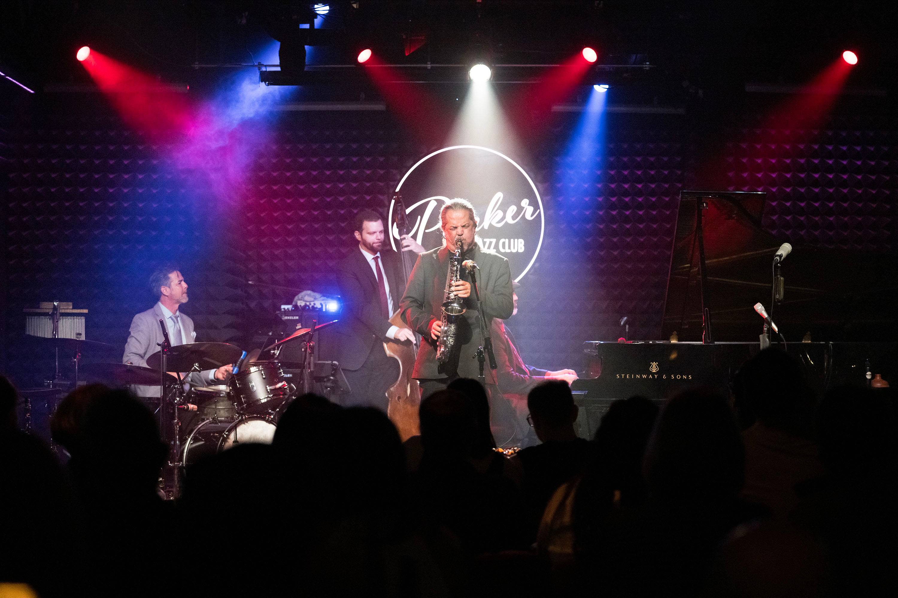 A group of musicians play various instruments on stage in a jazz club with red white and blue lights hitting them.