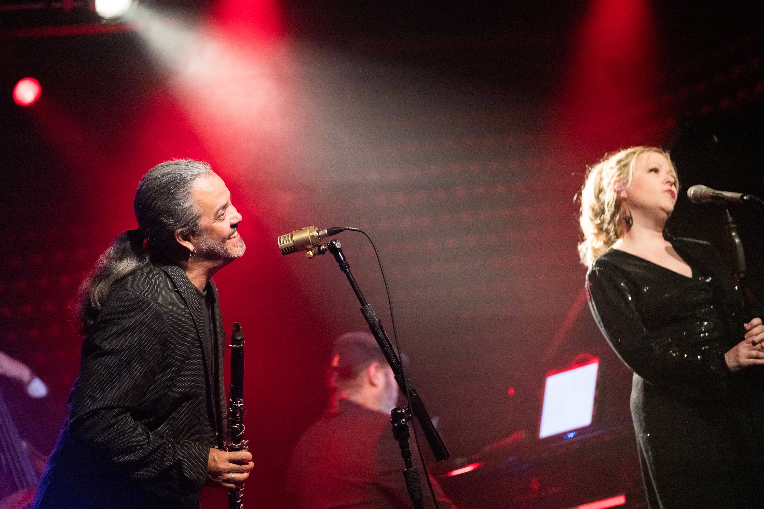 Man with a ponytail sings into a microphone on the left, on the right a blonde woman also sings into a microphone. The background is bathed in red light.